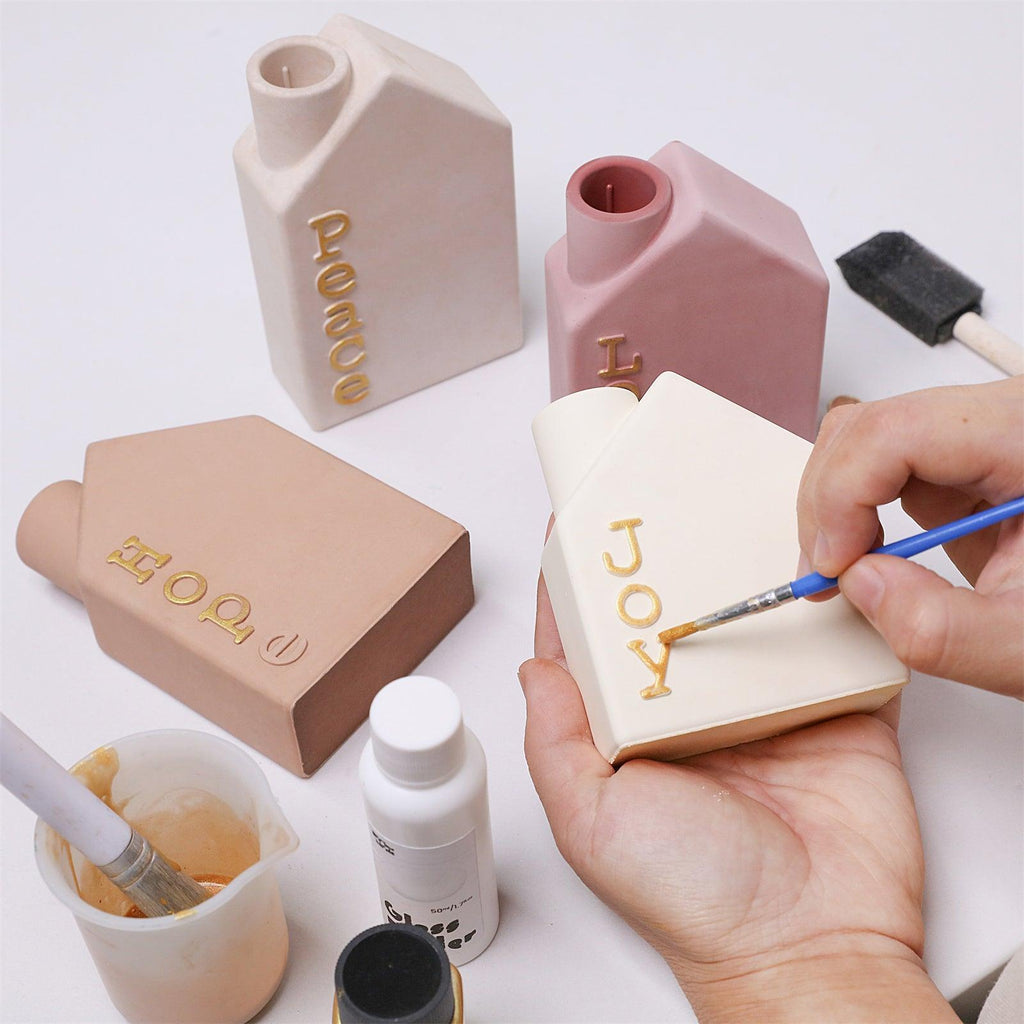 Hand painting gold letters on house-shaped Advent candle holder with “JOY” text - Boowan Nicole