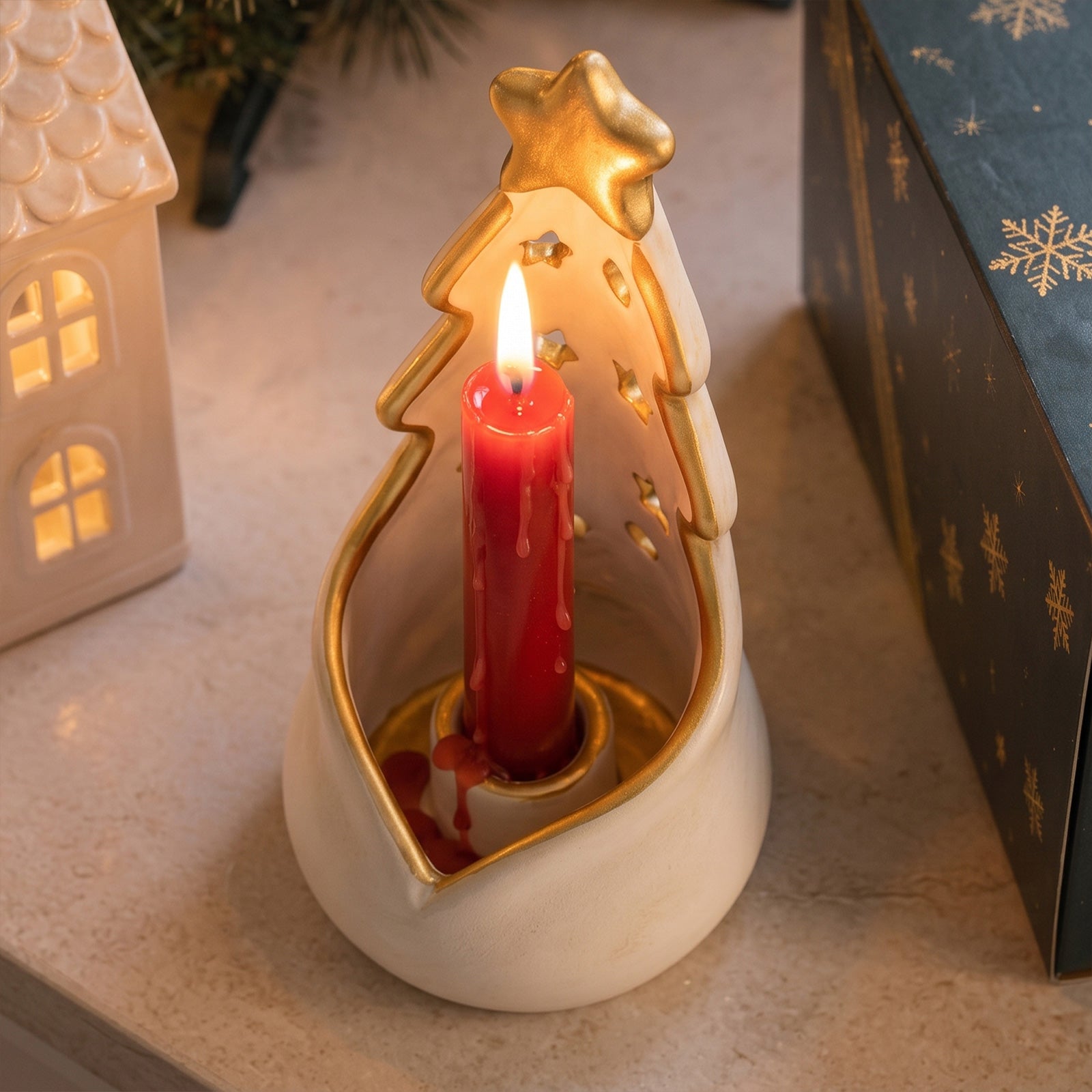 A Christmas tree-shaped white candle holder with gold trim, lit with a red pillar candle, set on a tabletop next to a small house-shaped ornament and a gift box.