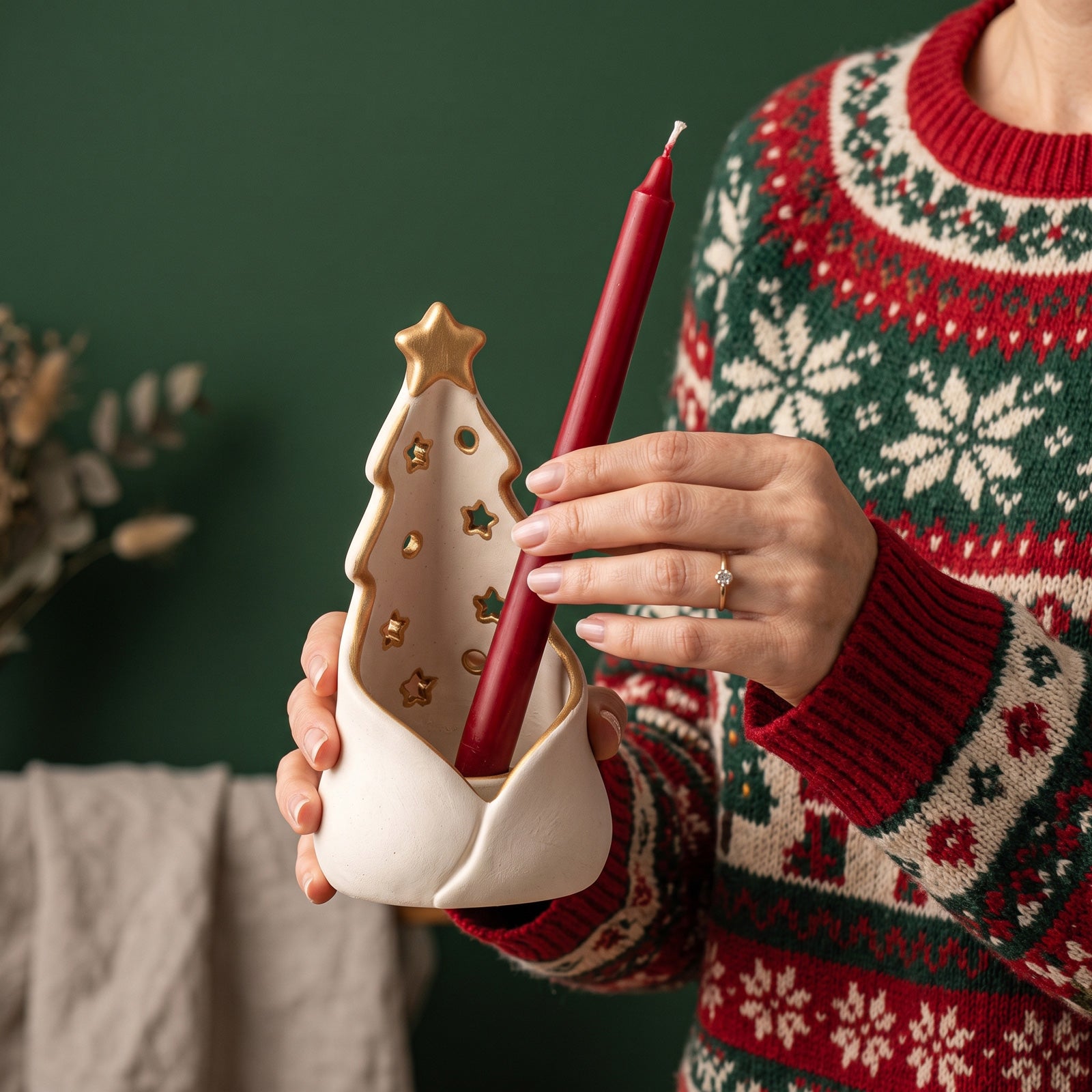 A person wearing a red, green, and cream Christmas sweater holds a red taper candle in a cream ceramic holder shaped like a Christmas tree with gold star details, set against a dark green background.