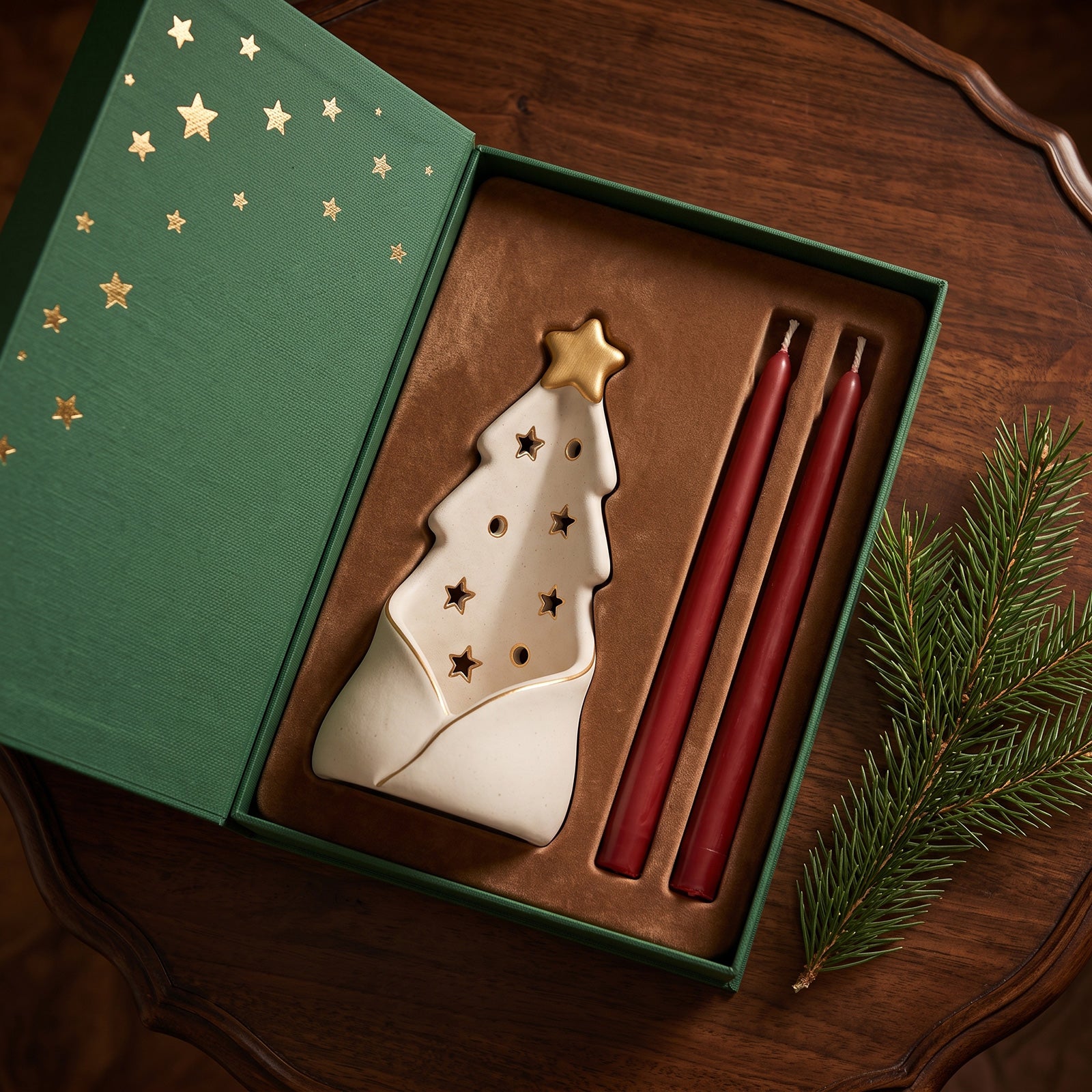 A Christmas-themed gift set in an open green box with gold stars, featuring a white ceramic tree-shaped candle holder with gold star cutouts and a gold star topper, alongside two red taper candles. The box is styled on a dark wooden table with a pine branch nearby.