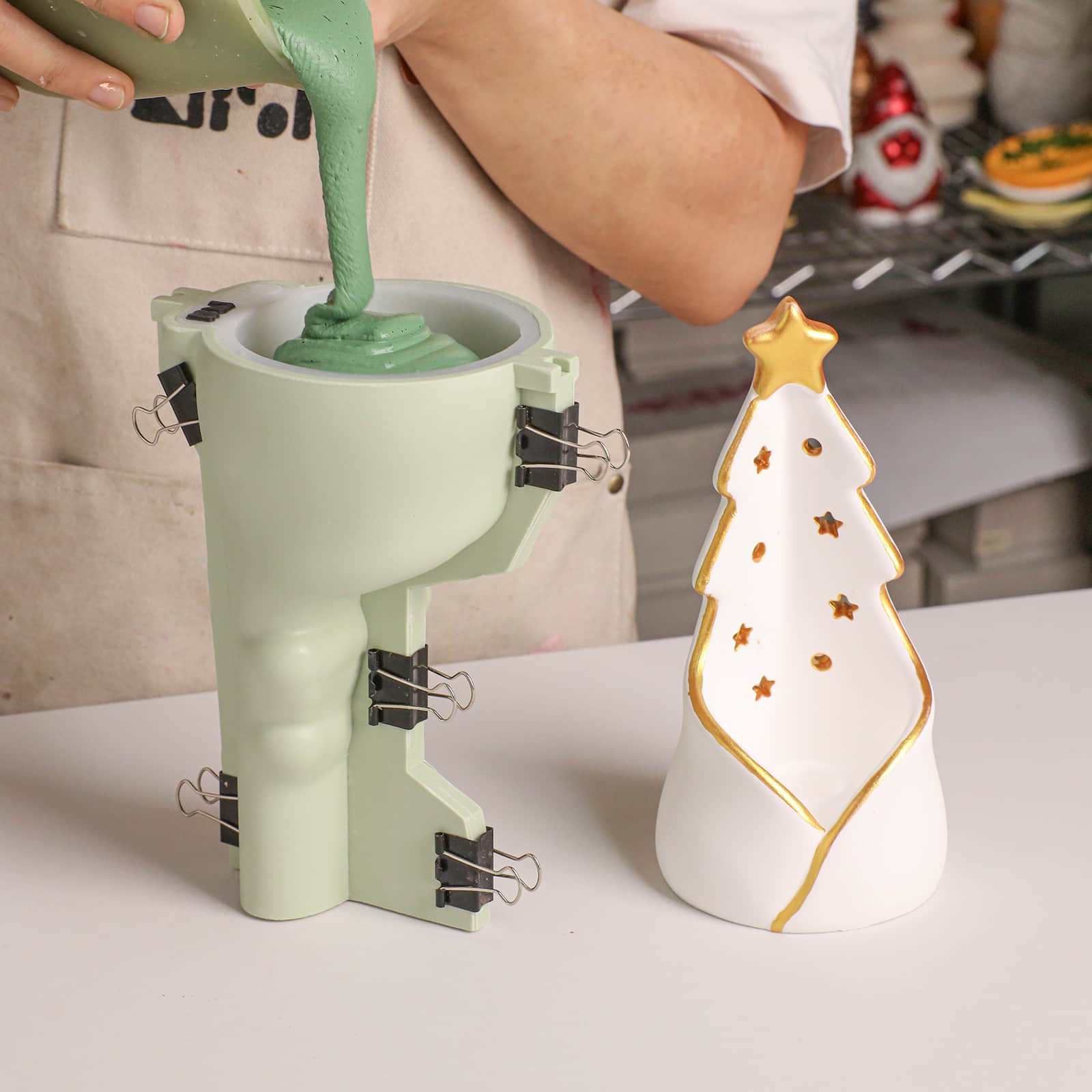 A person pours mint green silicone mold material into a pale green boot-shaped mold, with binder clips holding the mold open. A white Christmas tree-shaped ceramic candle or decor piece with gold trim sits beside the mold on a white tabletop.