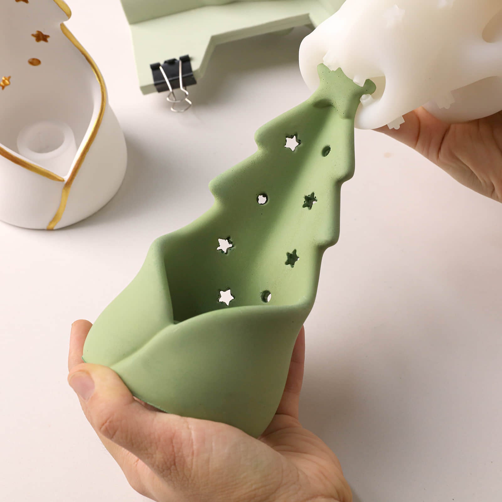 A person holds a light green silicone mold shaped like a Christmas tree with star-shaped cutouts, while pouring white silicone from another mold into it. The background is a clean white surface with other crafting items partially visible.