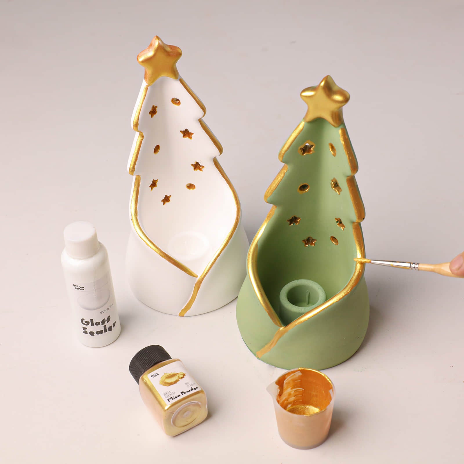 Two Christmas tree-shaped ceramic candle holders, one white and one sage green, both with gold metallic trim and star cutouts. A hand is holding a paintbrush applying gold paint to the green tree.