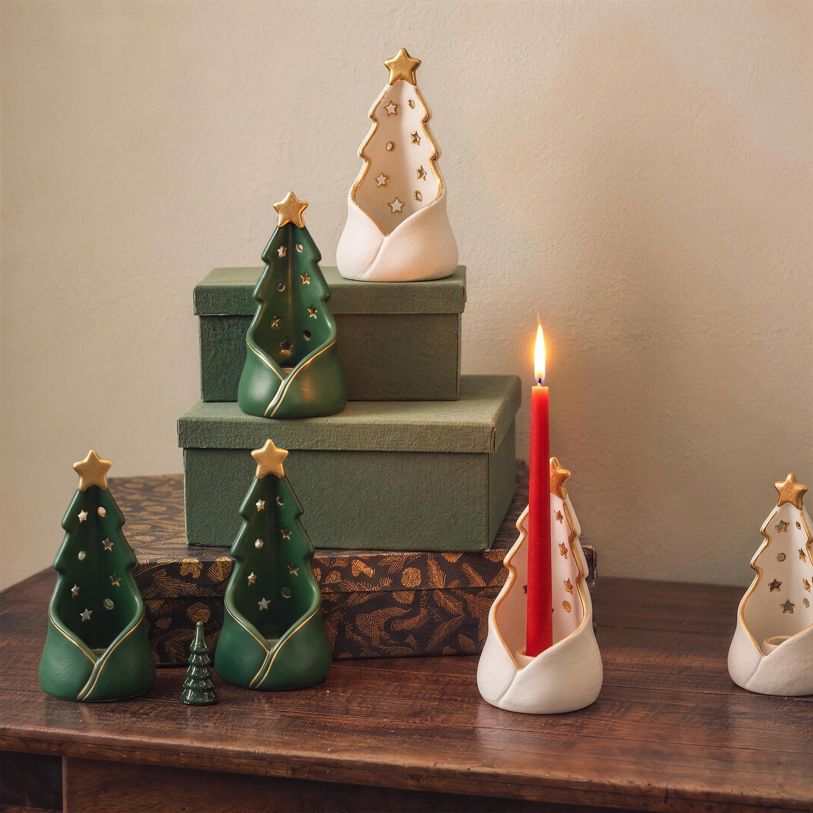 A set of ceramic Christmas tree-shaped candle holders in white and green, decorated with gold star and tree patterns, arranged on a wooden table with a lit red candle in one holder.