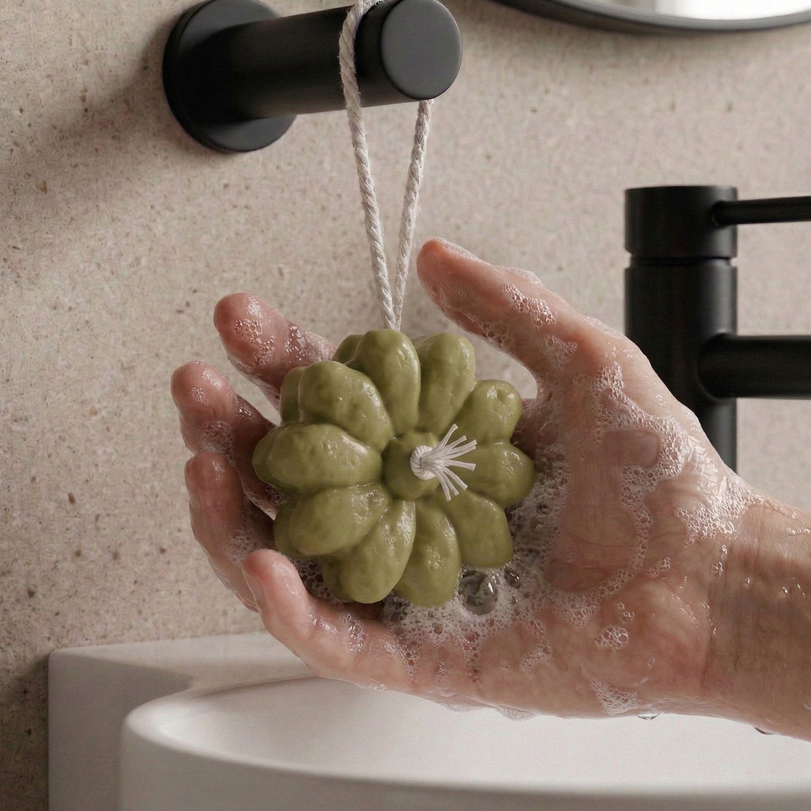 Hand holding a hanging green flower-shaped soap with lather near a bathroom faucet - Boowan Nicole