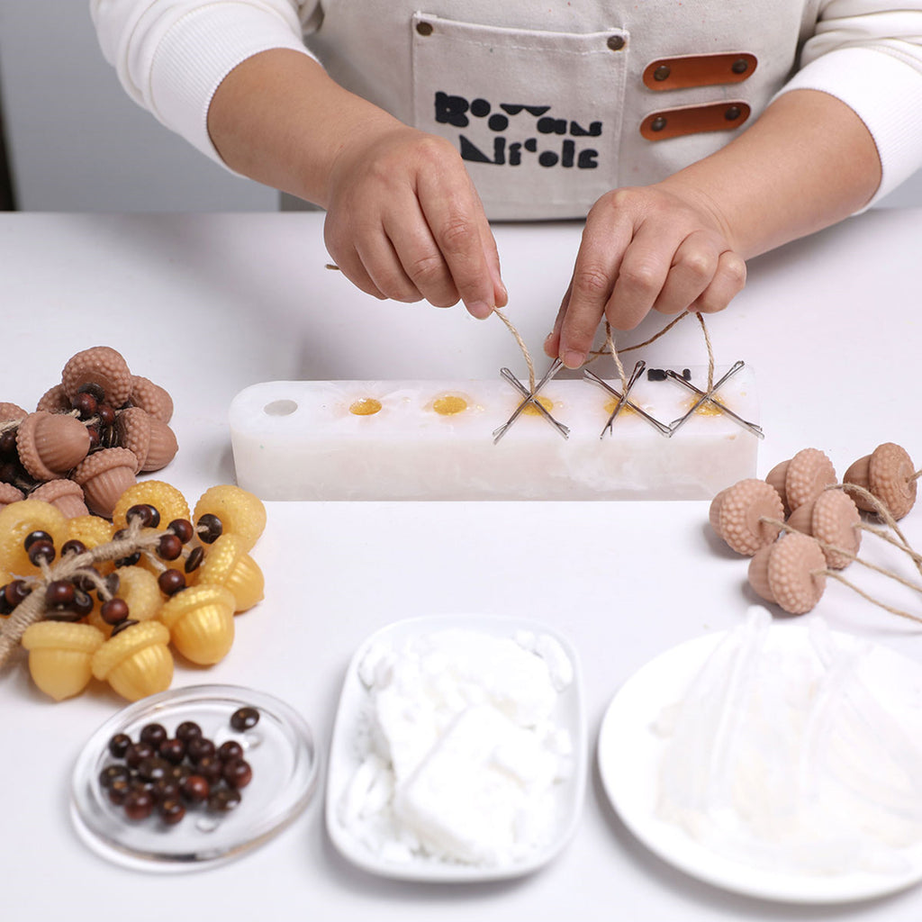 Hands threading jute through mini acorn-shaped soaps on a jig—making a hanging soap bunch with beads and supplies on the table - Boowan Nicole