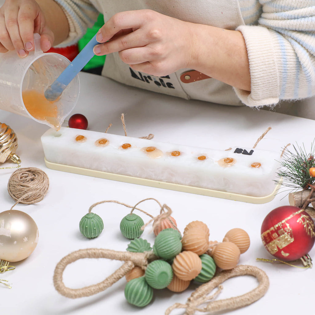 Pouring soap into a long silicone mold for hanging soap clusters; ribbed soaps and holiday ornaments nearby - Boowan Nicole