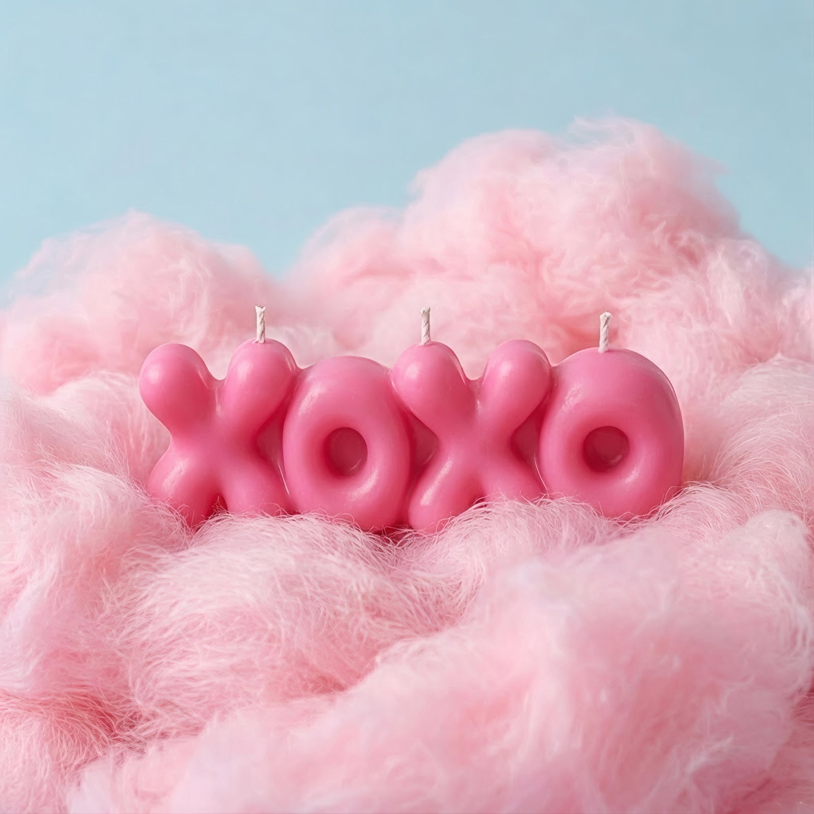 Bright pink "XOXO" bubble letter candle resting on fluffy pink faux fur, with a light blue backdrop. -Boowan Nicole