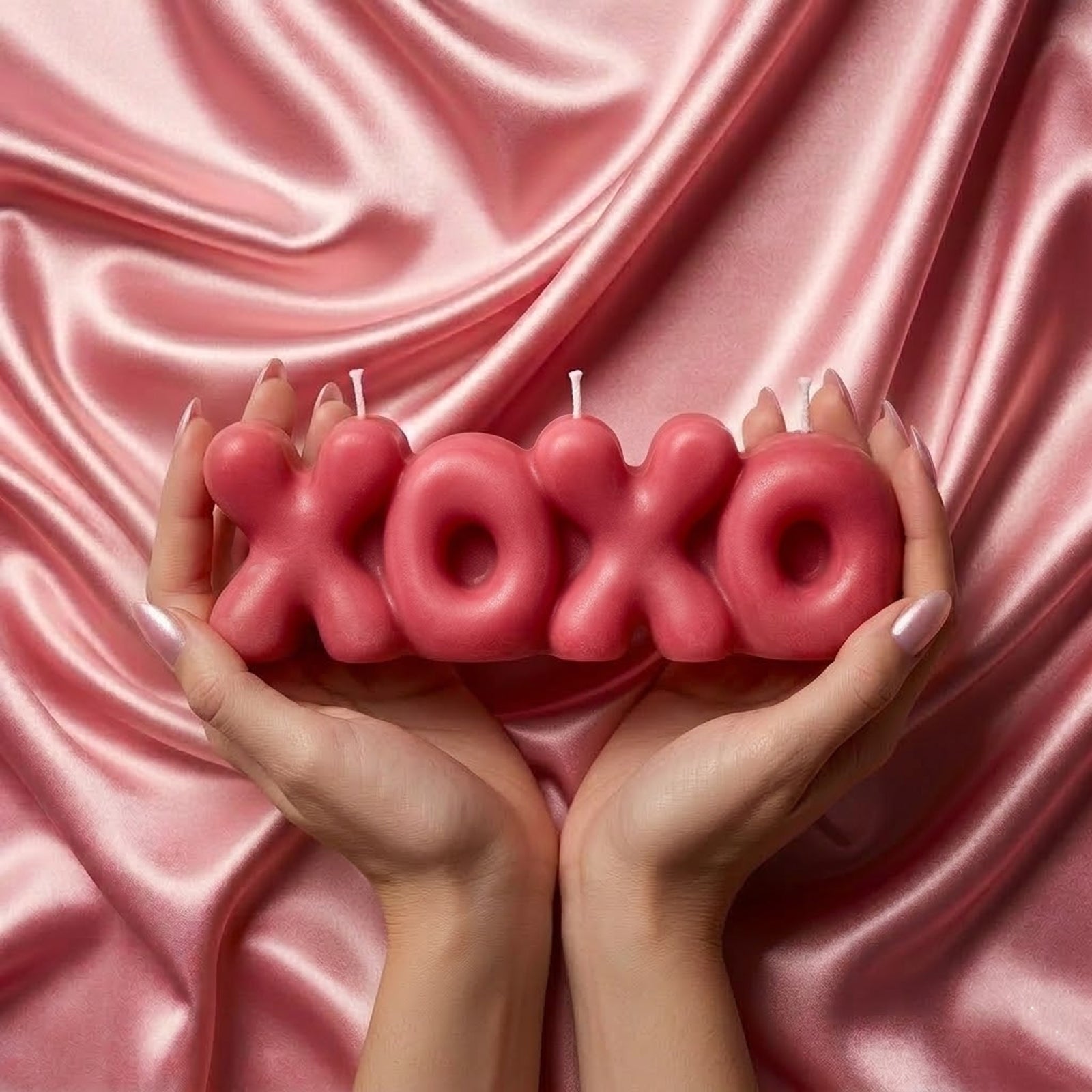 Two hands holding a deep pink "XOXO" bubble letter candle against glossy pink satin fabric. -Boowan Nicole