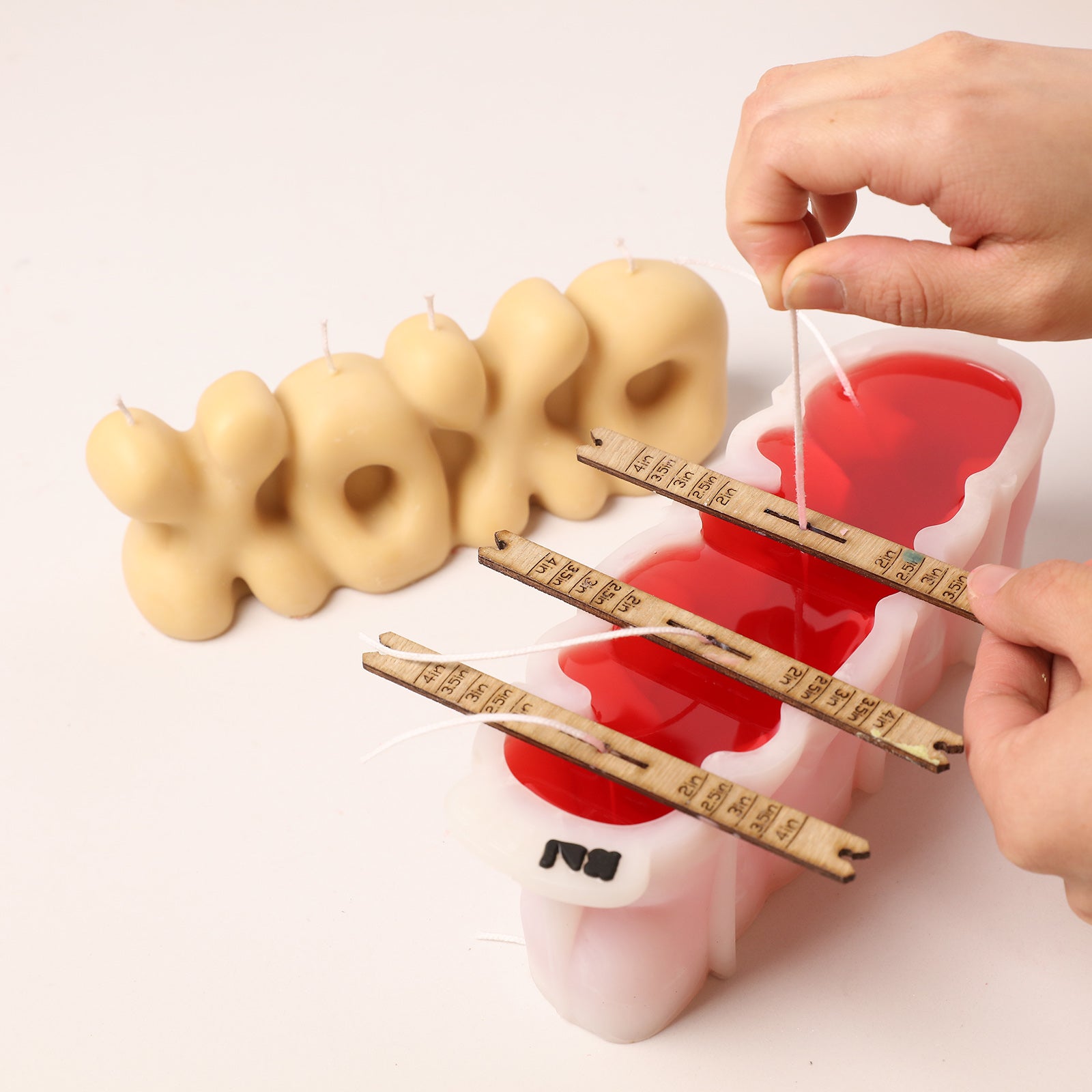 Crafting process: Placing wicks into a white silicone "XOXO" mold, with a finished beige "XOXO" candle displayed. -Boowan Nicole