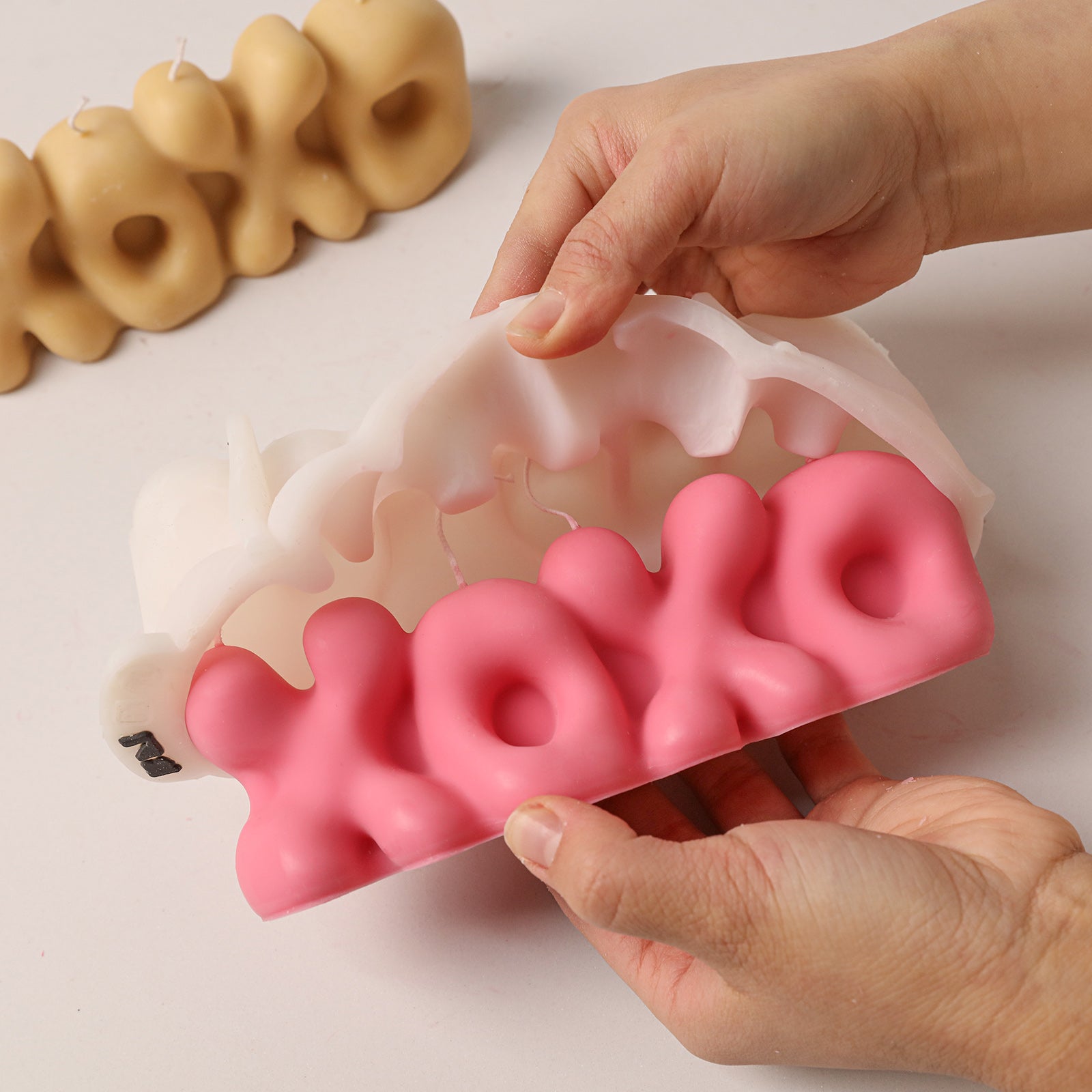 Demolding a bright pink "XOXO" bubble letter candle from a white silicone mold, with a beige candle in the background. -Boowan Nicole