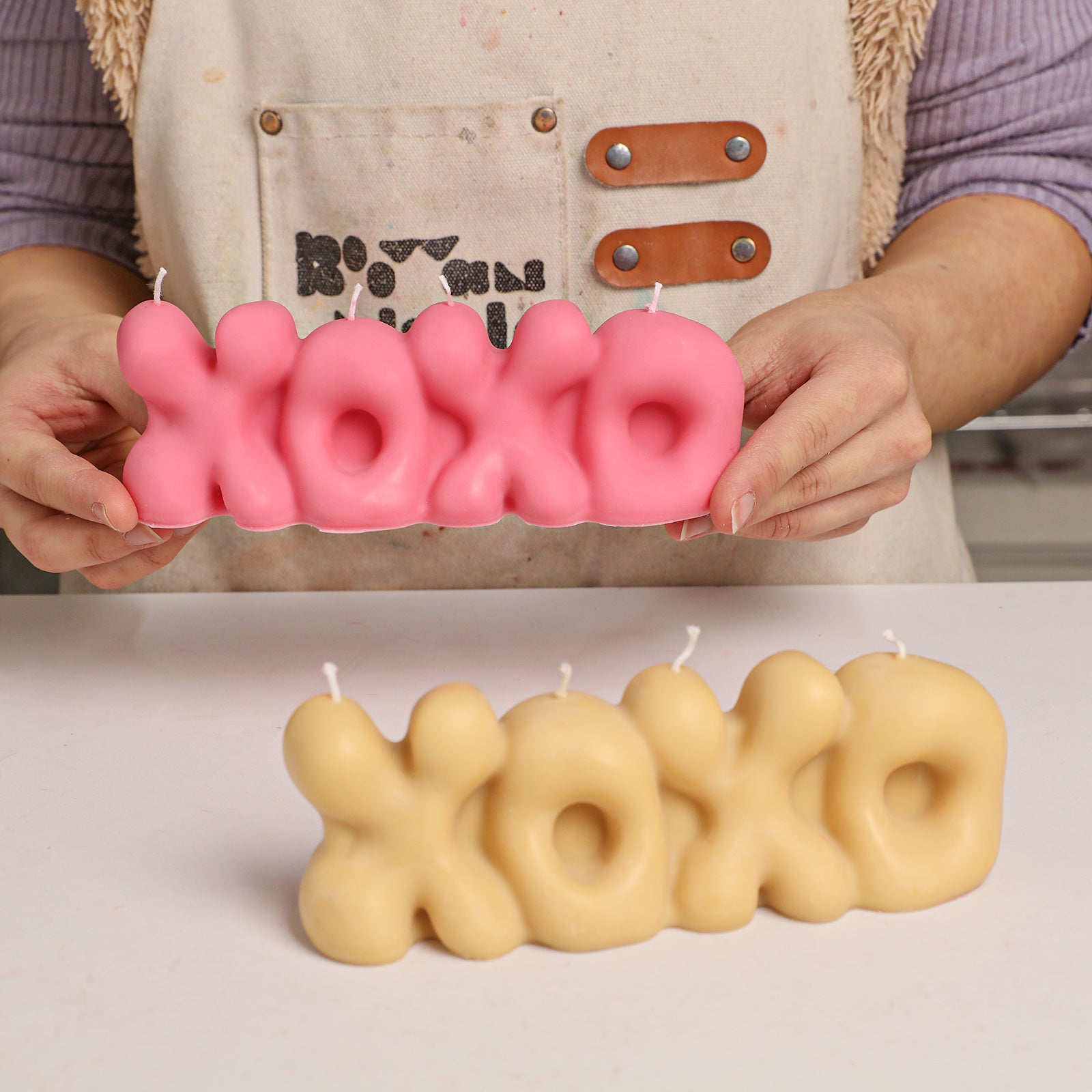 A person holding a bright pink "XOXO" bubble letter candle, with finished pink and beige "XOXO" candles on the table. -Boowan Nicole