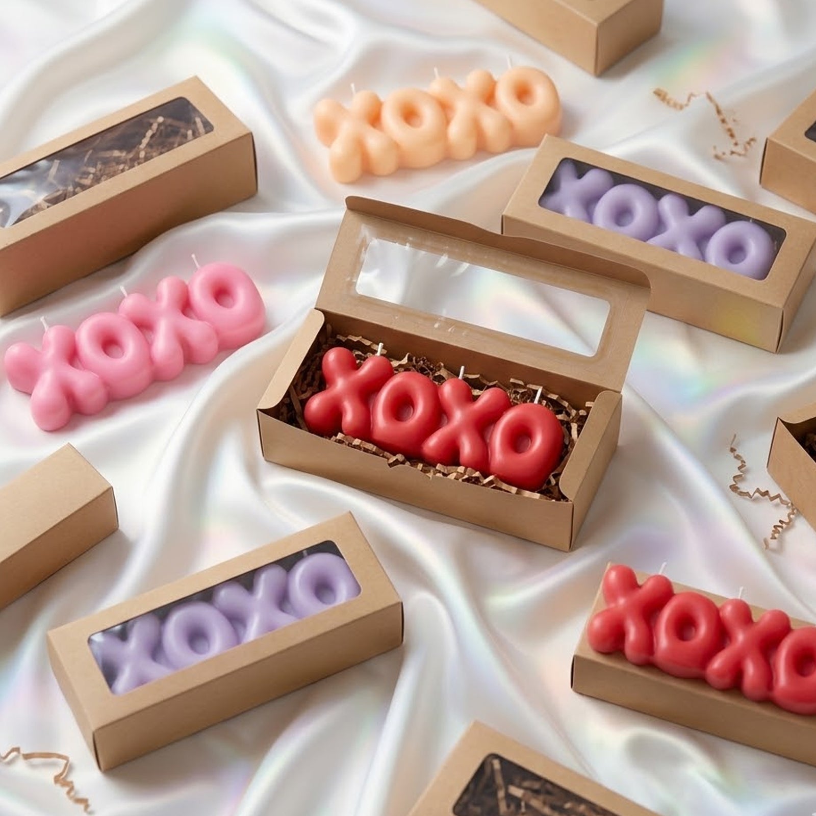 Colorful "XOXO" bubble letter candles (pink, red, peach, purple) in kraft gift boxes, arranged on iridescent white satin. -Boowan Nicole
