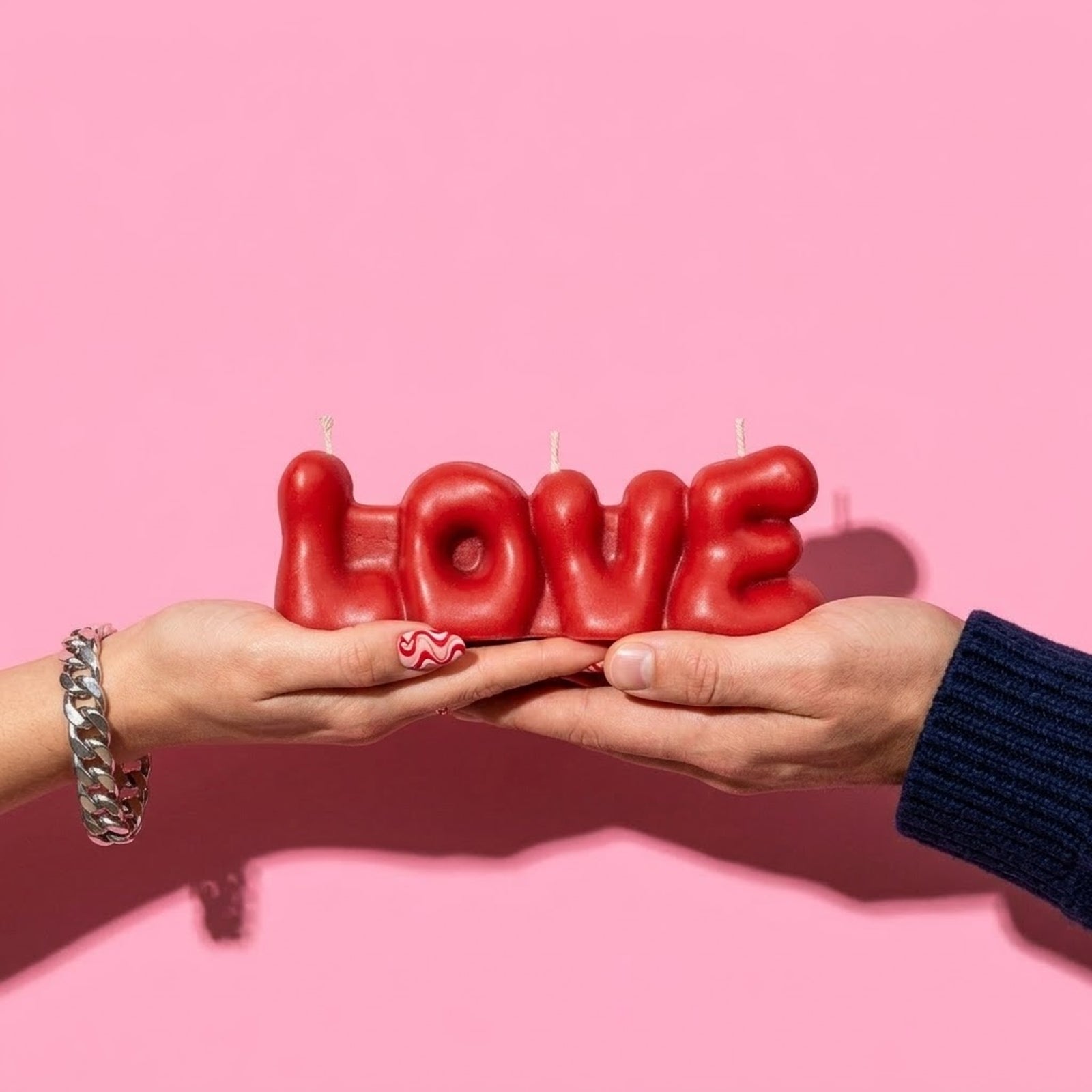 Two hands holding a red "LOVE" bubble letter candle, styled against a light pink background. -Boowan Nicole
