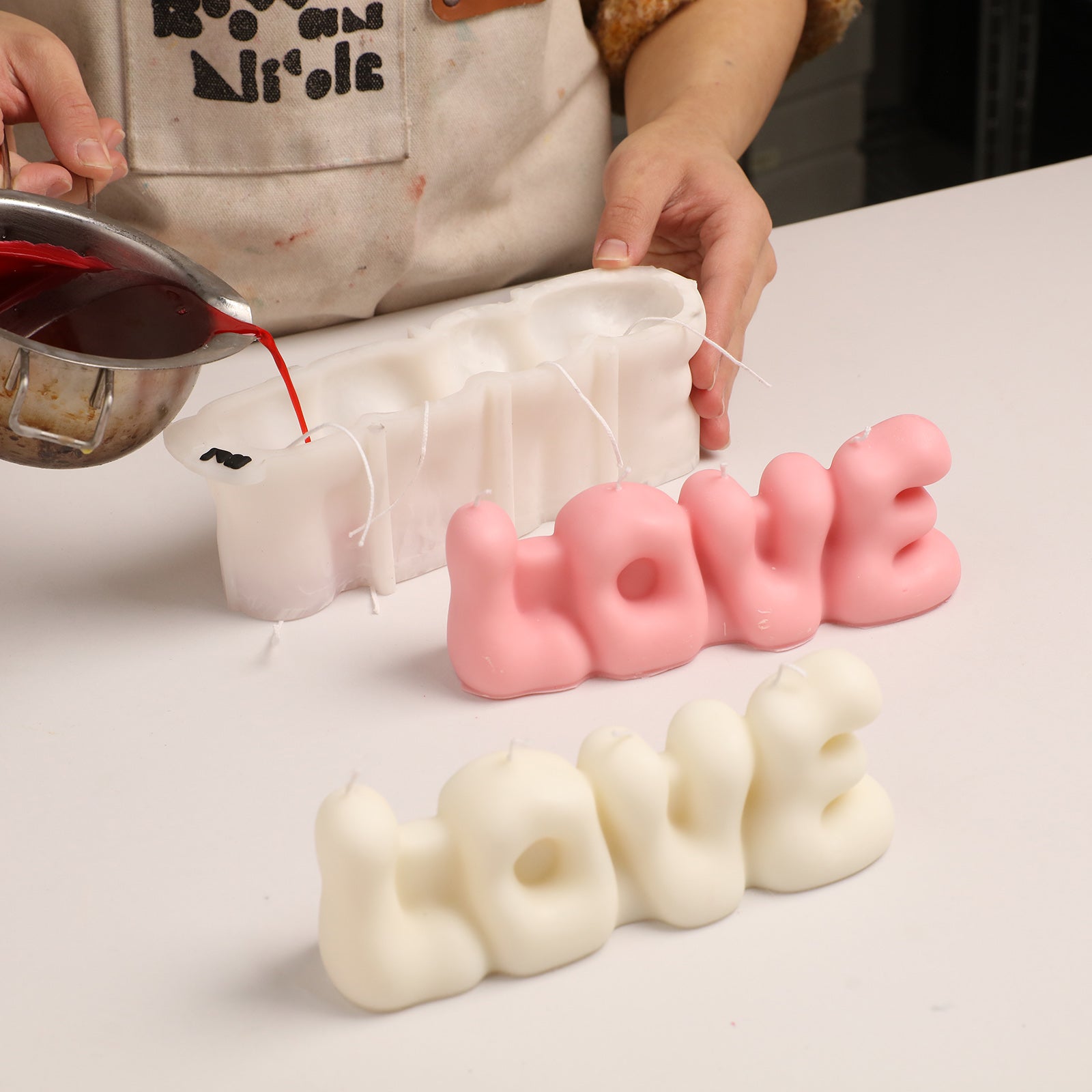 Crafting process: Pouring red wax into a "LOVE" bubble letter silicone mold, with finished pink and white "LOVE" candles displayed. -Boowan Nicole