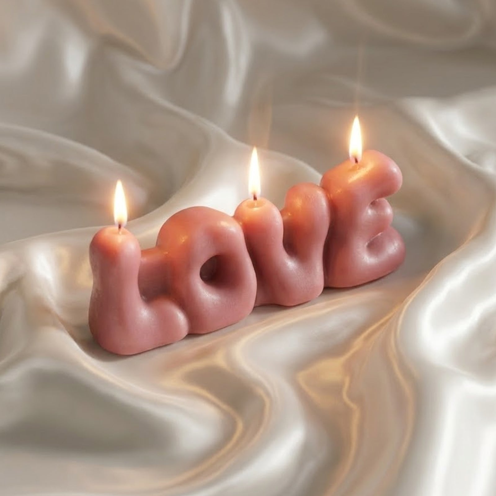 Lit dusty pink "LOVE" bubble letter candle resting on white satin fabric, creating a soft, romantic glow. -Boowan Nicole