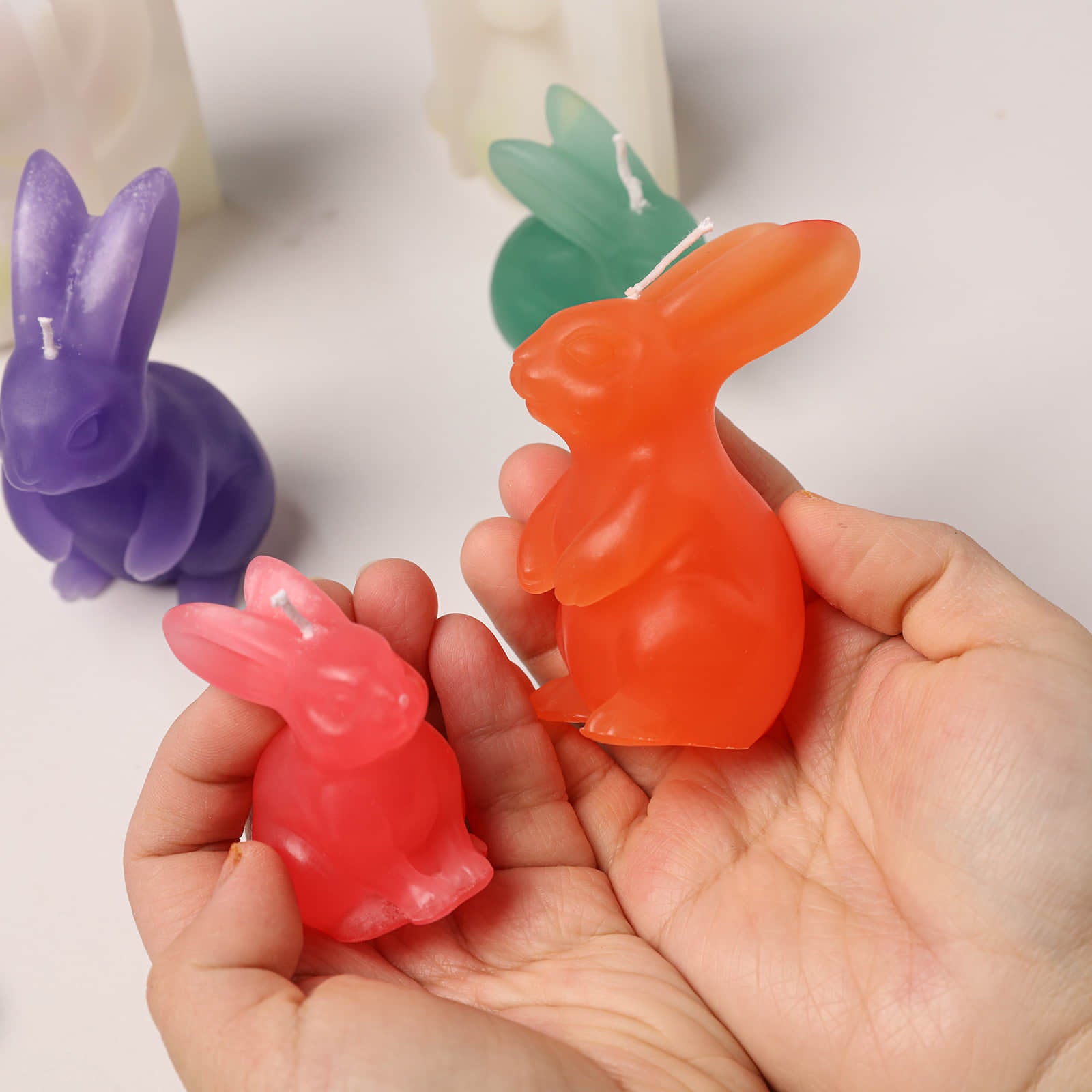 Hand holding pink and orange Bunny Buddy candles; colorful rabbit-shaped candles and molds in the background - Boowan Nicole