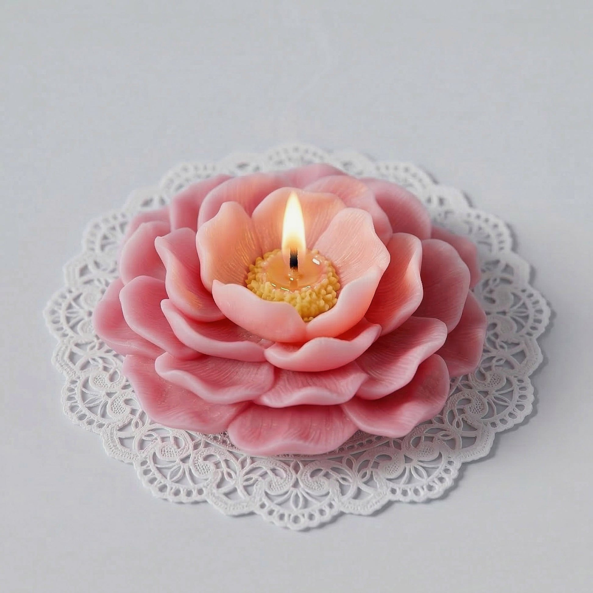 A lit pink camellia flower candle resting on a white lace doily, casting warm glow. -Boowan Nicole