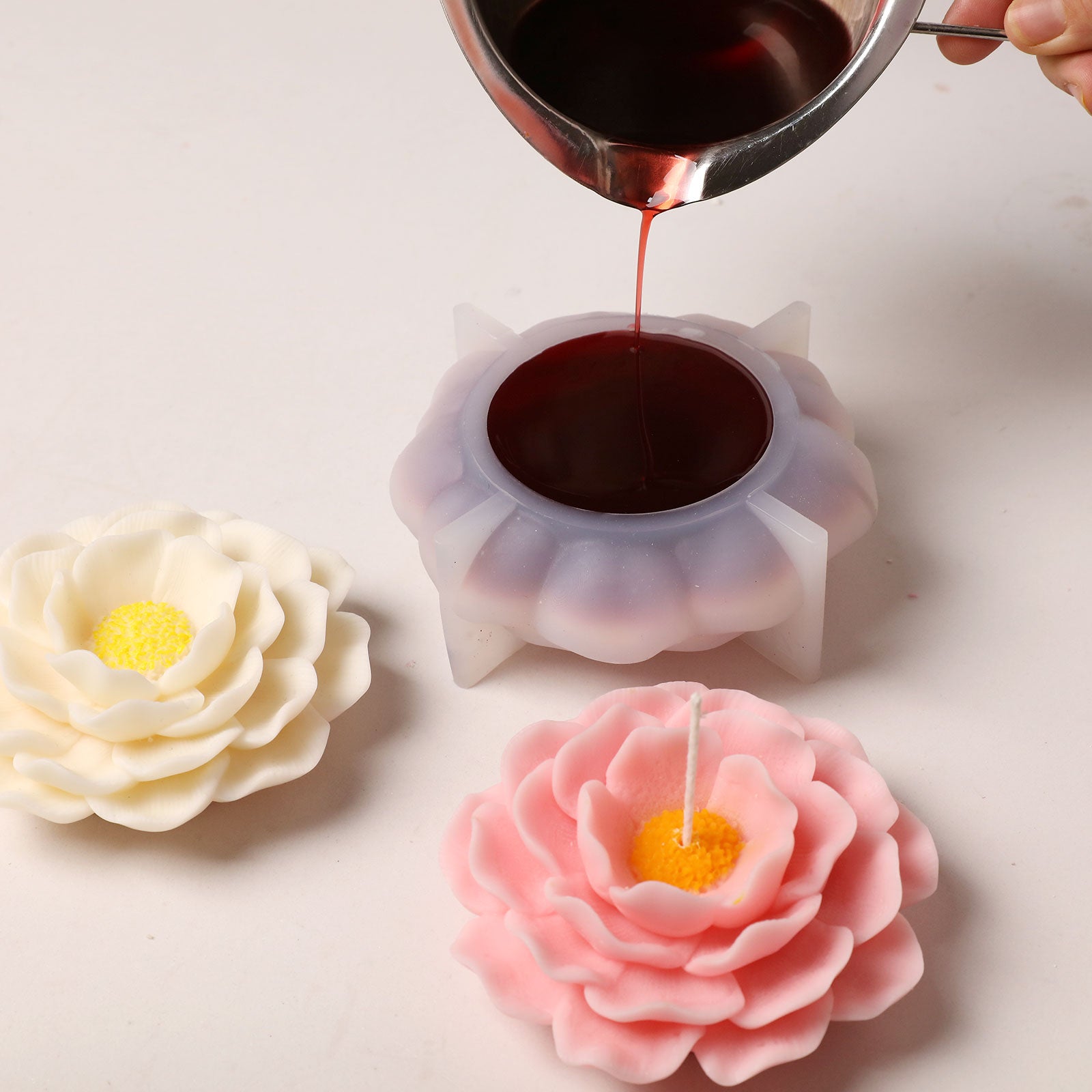 Pouring deep red wax into a camellia-shaped silicone mold, with finished white and pink candles displayed. -Boowan Nicole