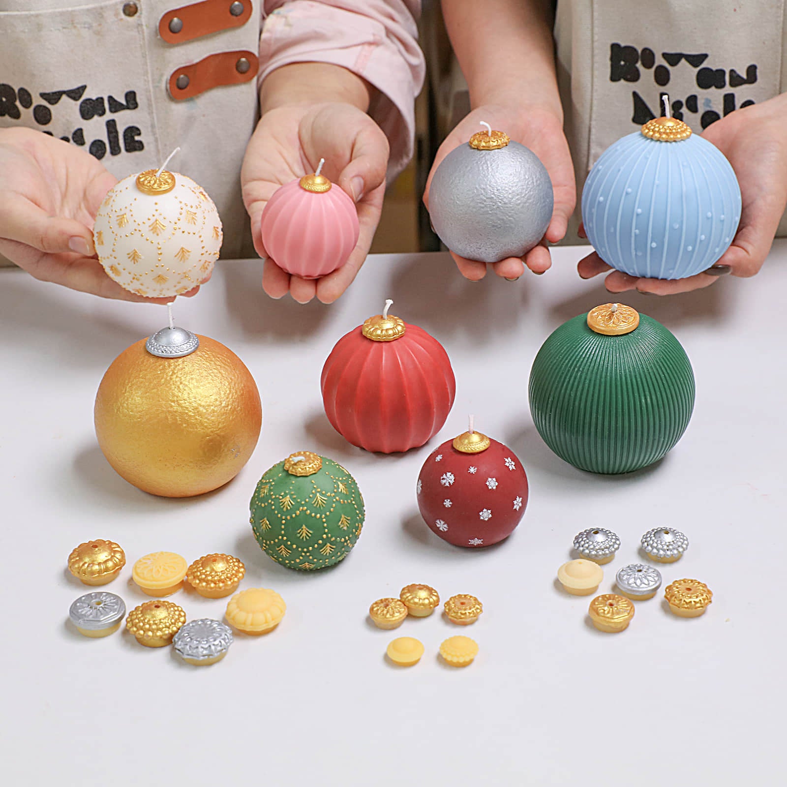 Hands displaying assorted bauble candles—gold hammered, pink fluted, green ribbed, blue pinstripe-dot, silver, white lace, red snowflake—with gold/silver caps and topper pieces on the table - Boowan Nicole