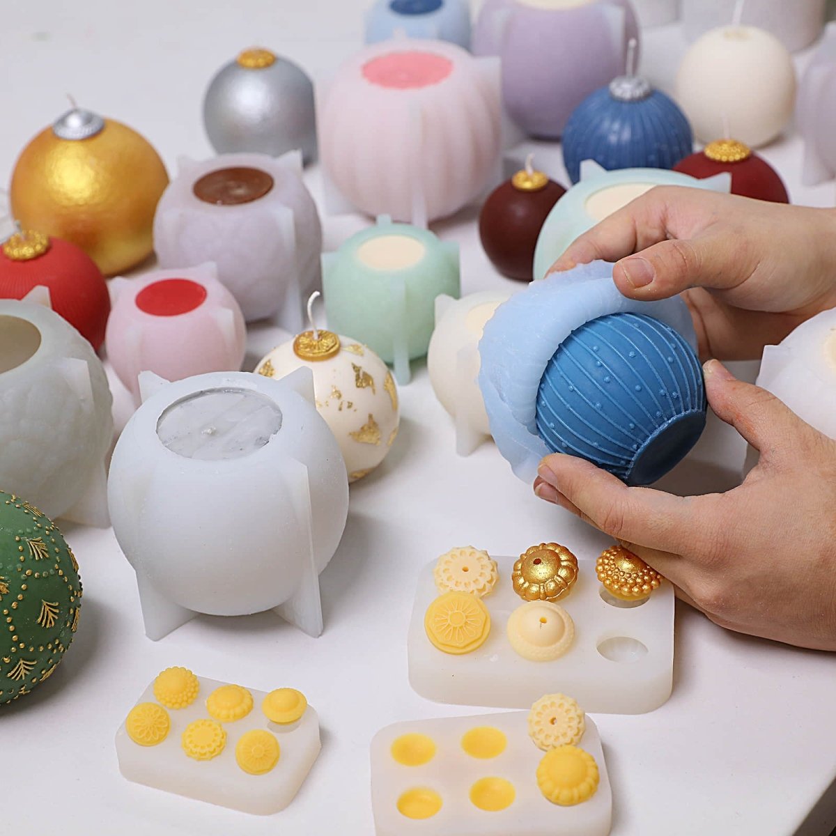 Hands demolding a blue pinstripe-dot bauble candle from a silicone mold, with finished candles, molds, and gold/silver topper caps on the table - Boowan Nicole