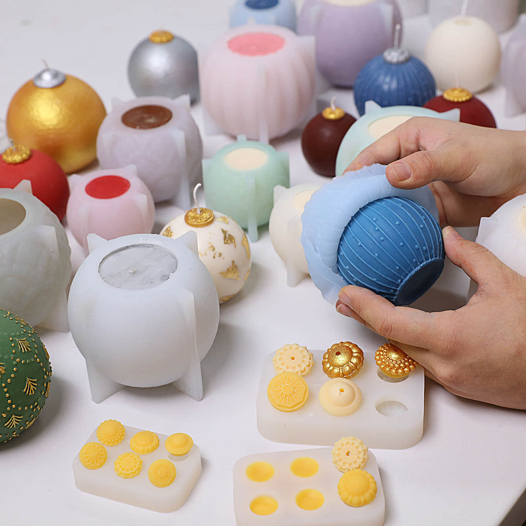 Hands demolding a blue pinstripe-dot bauble candle from a silicone mold, with finished candles, molds, and gold/silver topper caps on the table - Boowan Nicole