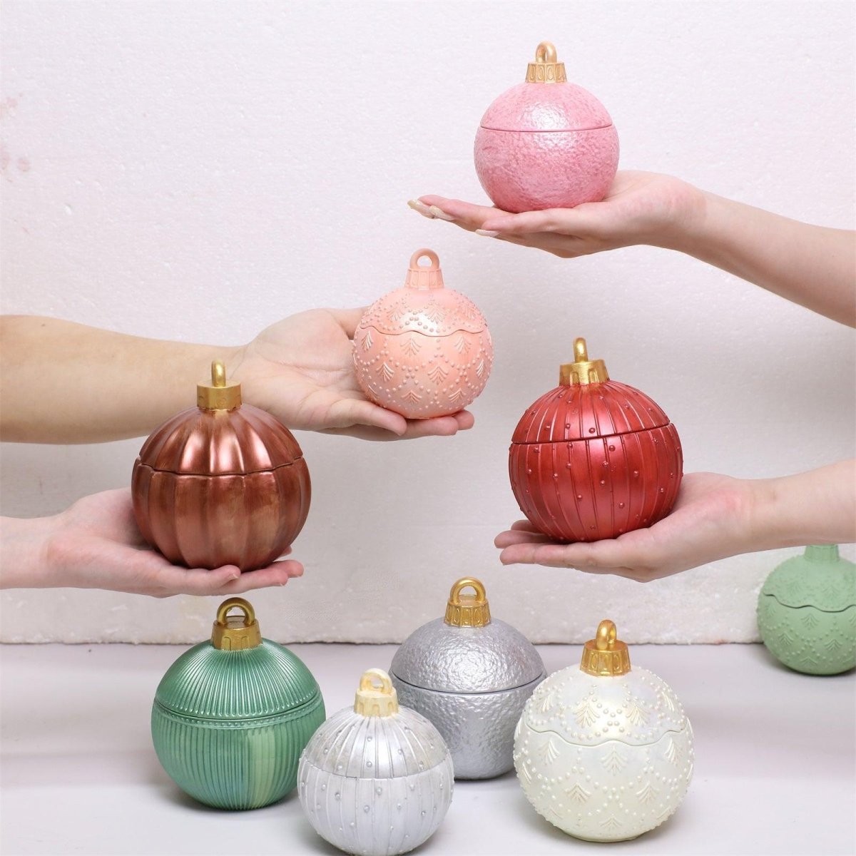 Hands holding colorful Christmas bauble candle jars in red, pink, and bronze, with additional green, silver, and white bauble jars displayed below - Boowan Nicole