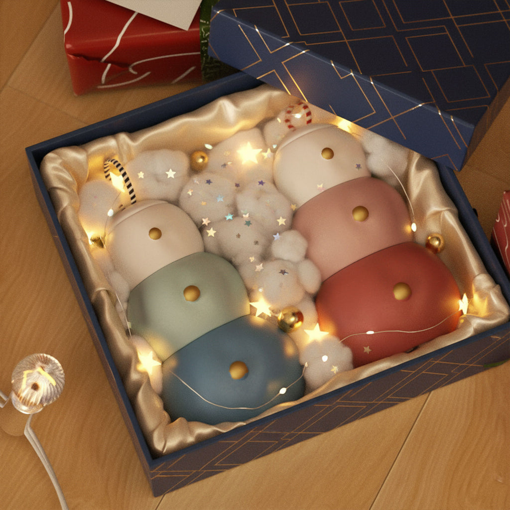 Gift box with two stacked snowman candle jars—blue-green and red-pink—featuring ring-top lids and gold button details, nestled in satin with fairy lights and star confetti - Boowan Nicole