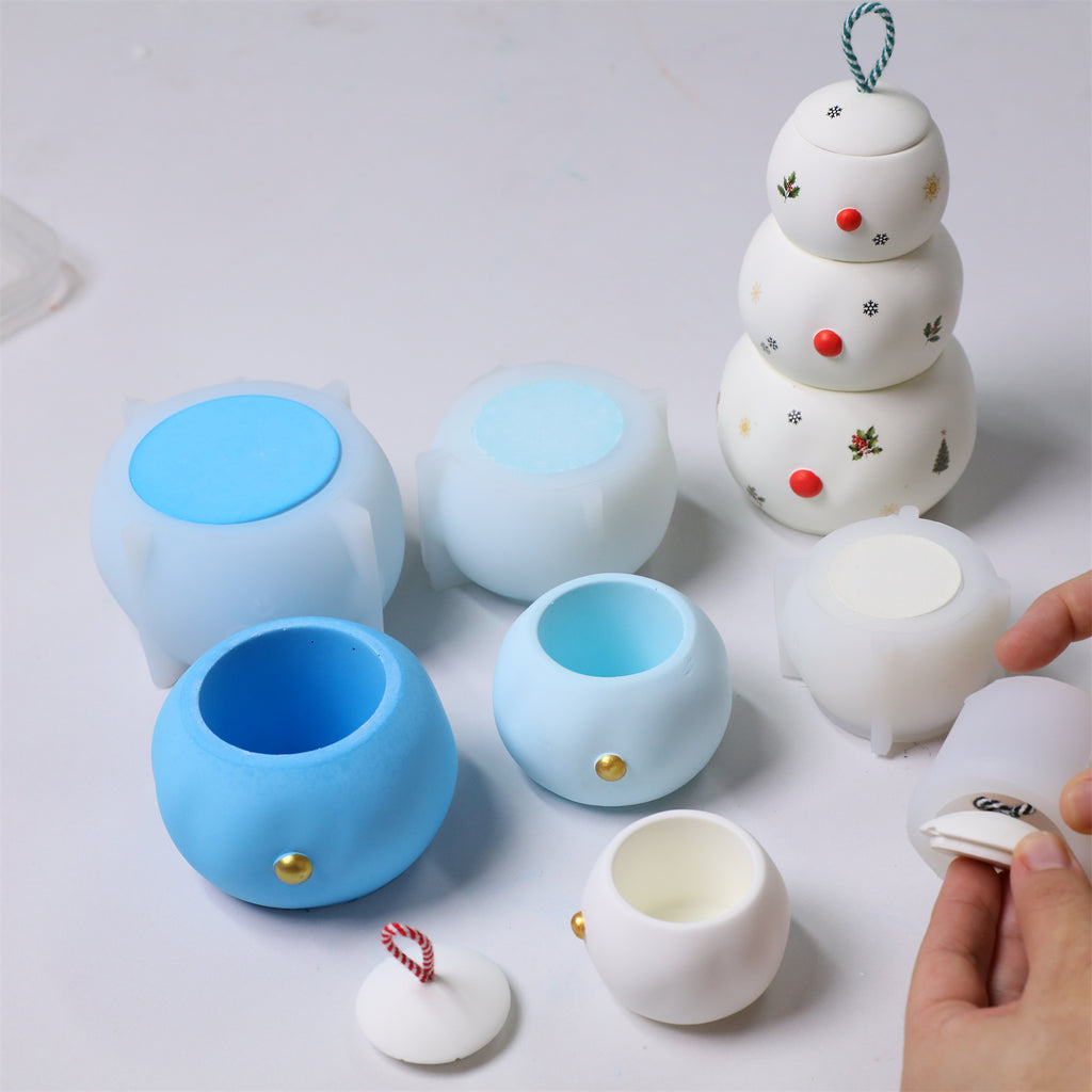 Snowman candle jar workshop—blue silicone molds and finished white jars with gold button details, candy-cane lid, and hands assembling parts - Boowan Nicole