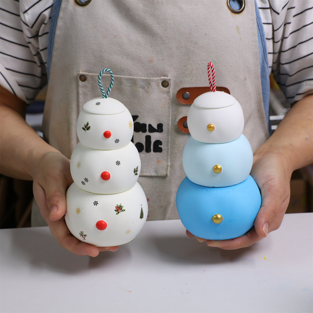 Two stacked snowman candle jars held in hands—white with holiday decals and blue gradient—ring-top lids with gold/red button details, maker’s apron backdrop - Boowan Nicole
