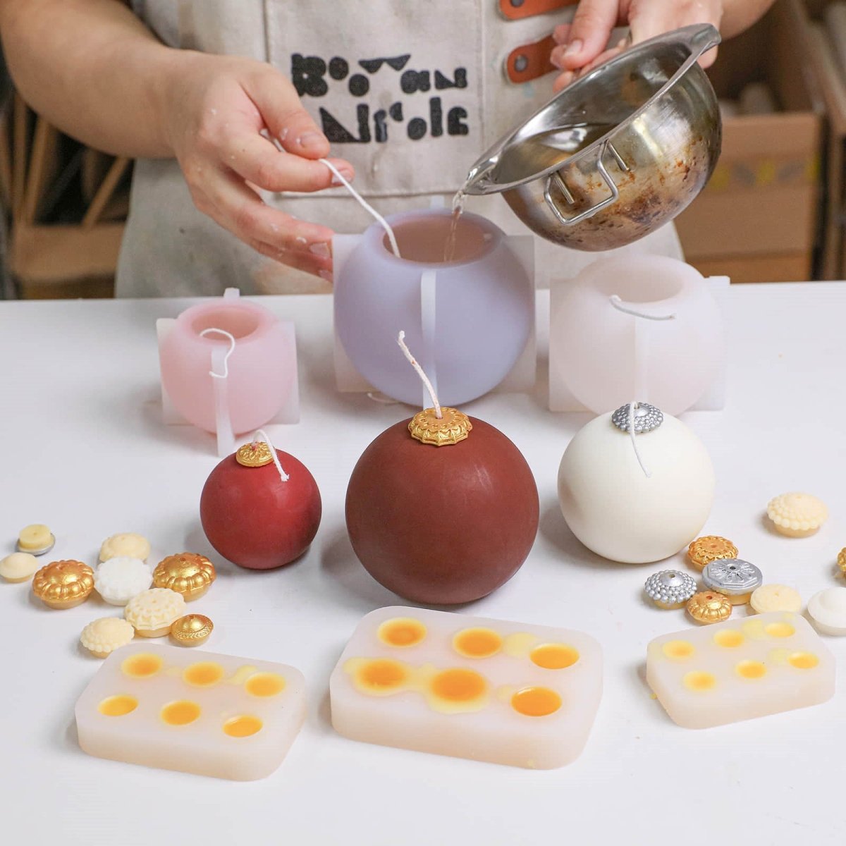 Hand pouring wax into smooth bauble candle silicone molds; finished glossy red and ivory candles with gold/silver caps and topper-cap molds on the table - Boowan Nicole