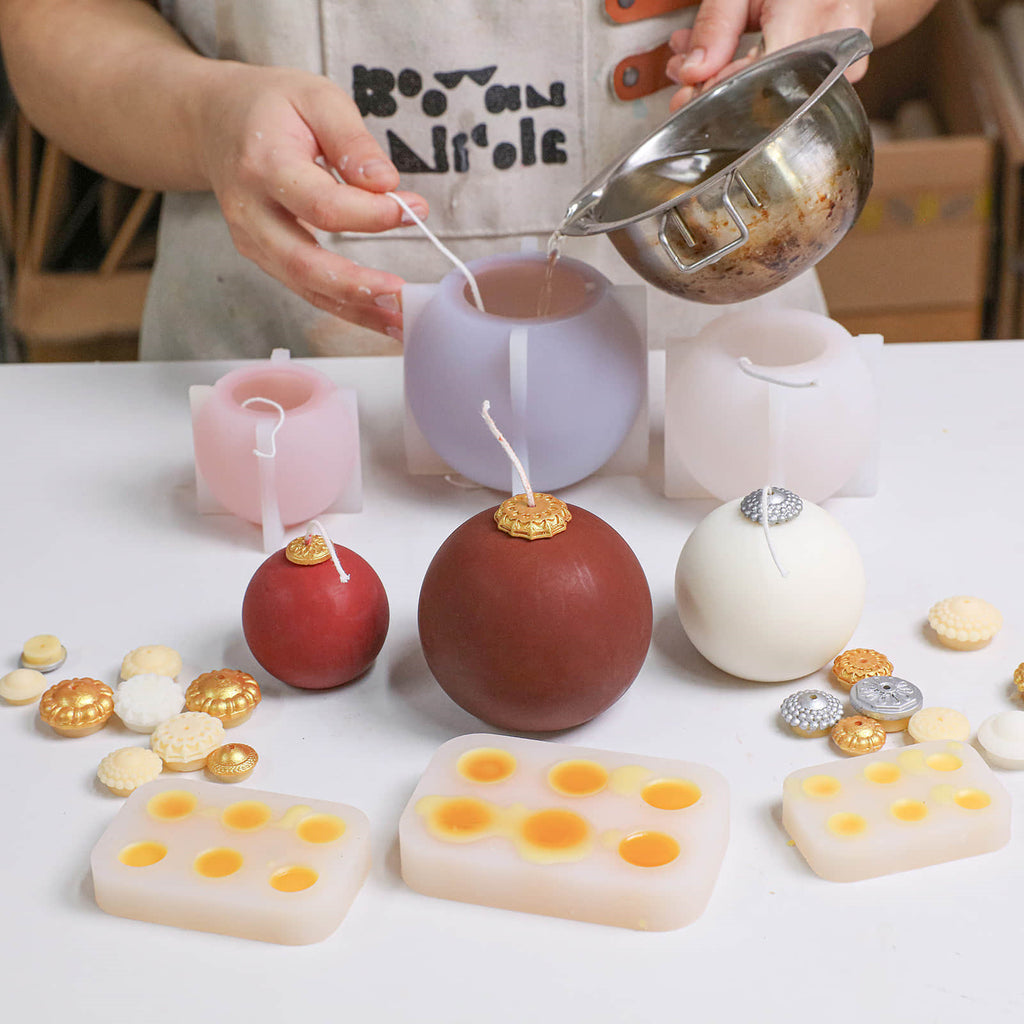 Hand pouring wax into smooth bauble candle silicone molds; finished glossy red and ivory candles with gold/silver caps and topper-cap molds on the table - Boowan Nicole