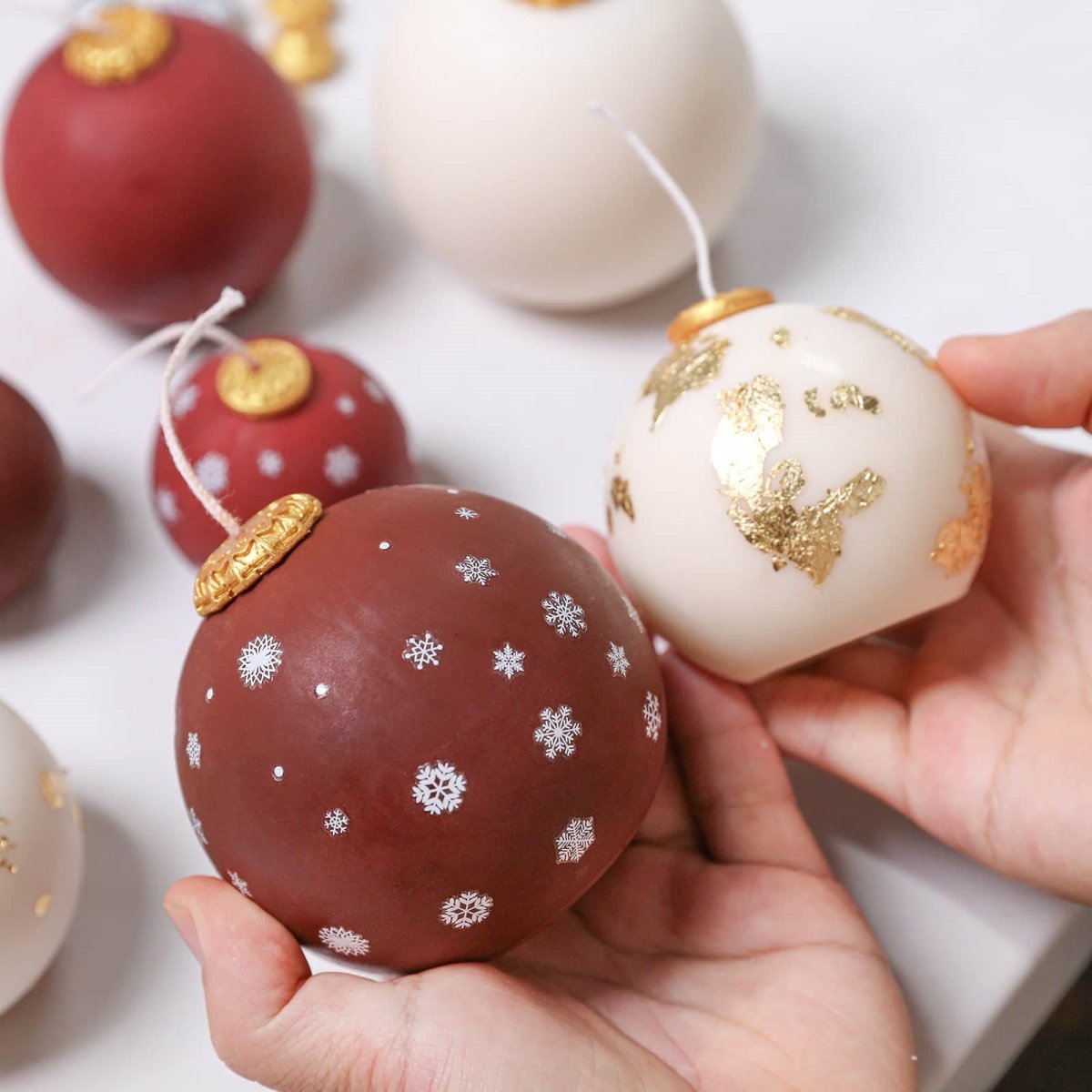 Hands holding cocoa-red bauble candle with silver snowflakes and ivory bauble with gold leaf, both with gold caps—DIY Christmas candles - Boowan Nicole