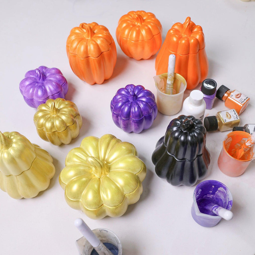 Hand-painted pumpkin candle jars in vibrant Halloween colors—gold, purple, black, and orange—displayed with mica varnishes and brushes - Boowan Nicole