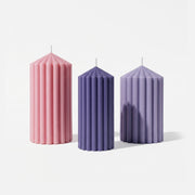 Three unlit ribbed pillar candles in pink, dark purple and light purple against a white background - Boowan Nicole