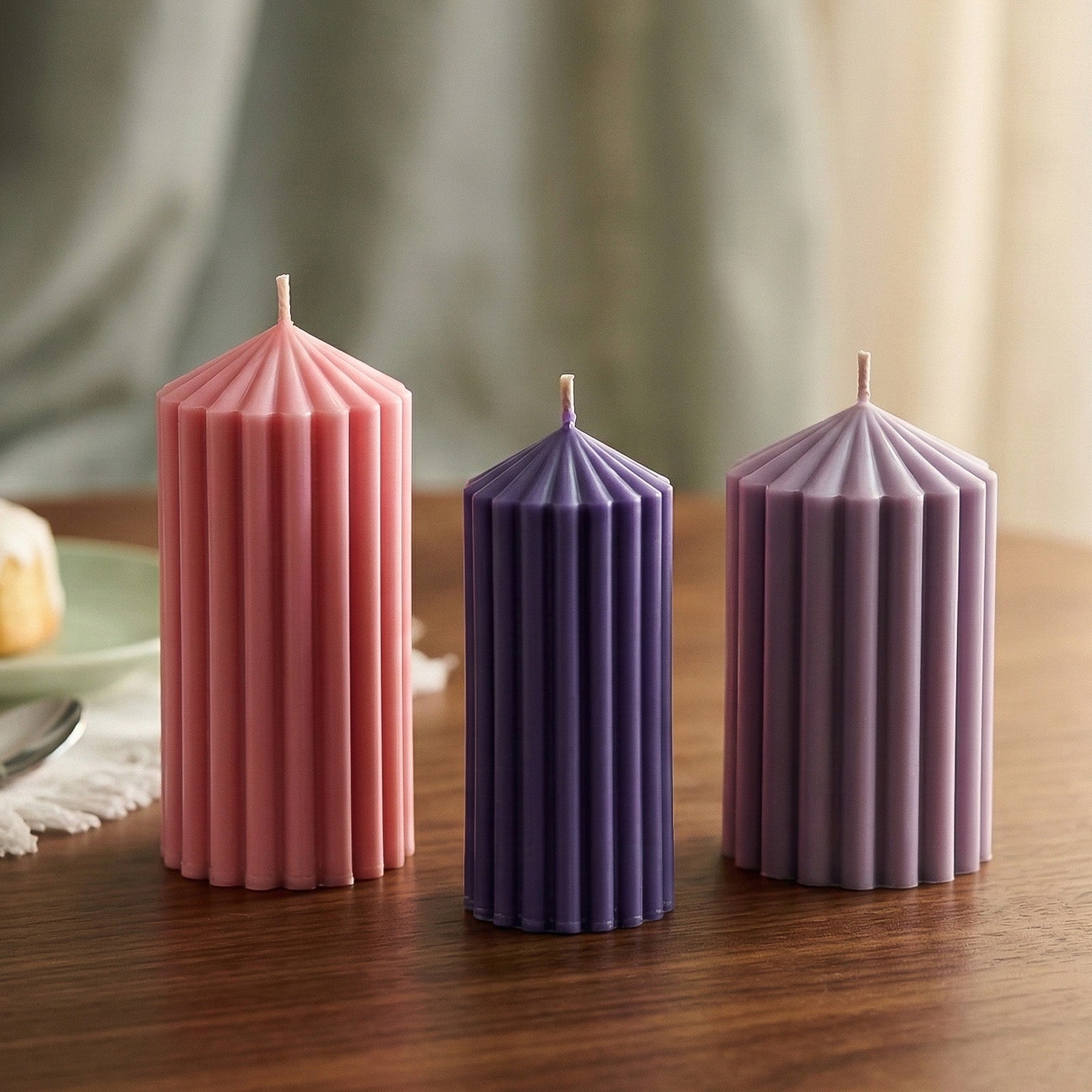 Three unlit ribbed pillar candles in pink, dark purple and light purple on a wooden table with a soft background - Boowan Nicole