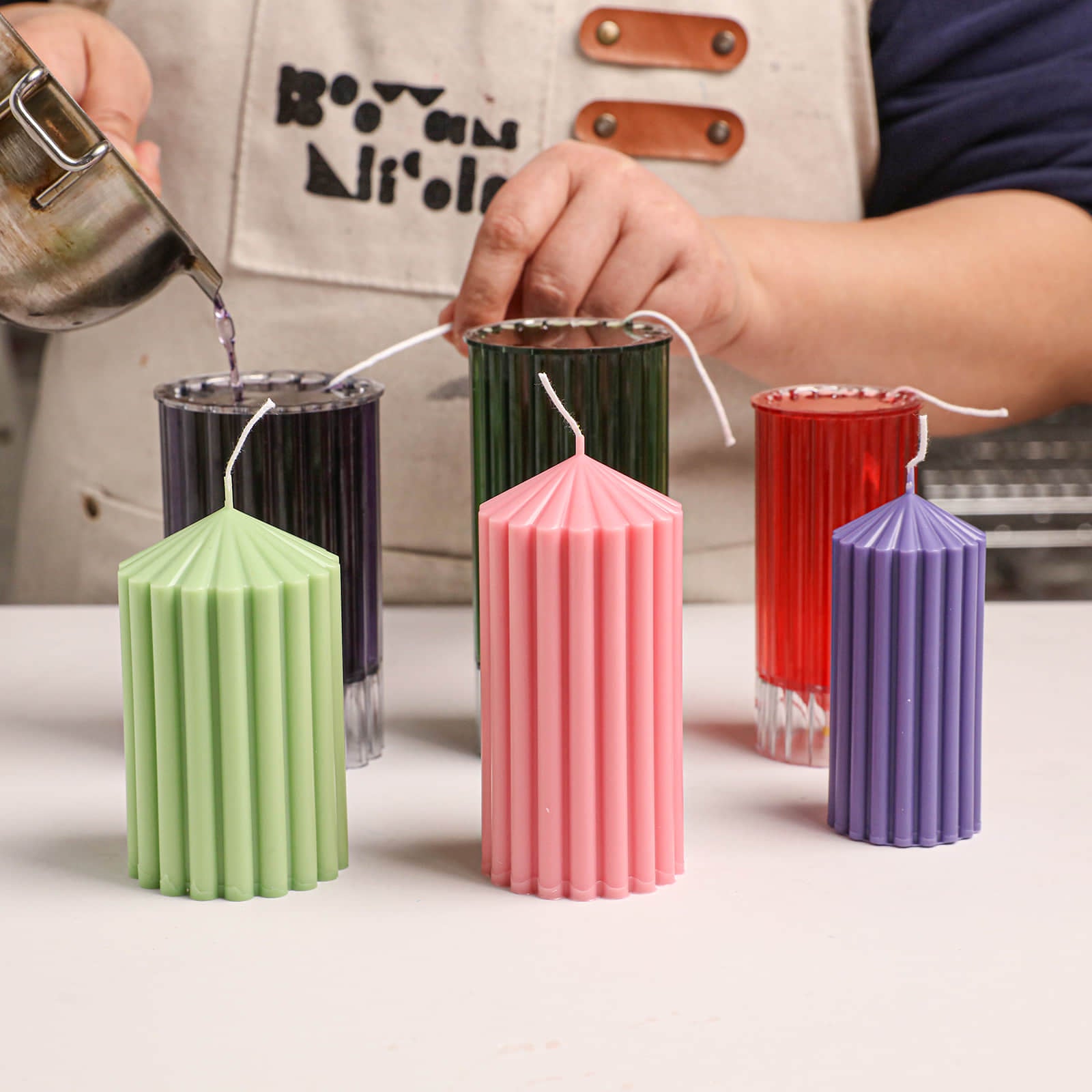 Candle making process: pouring wax into ribbed pillar candle acrylic molds with finished colorful candles nearby - Boowan Nicole