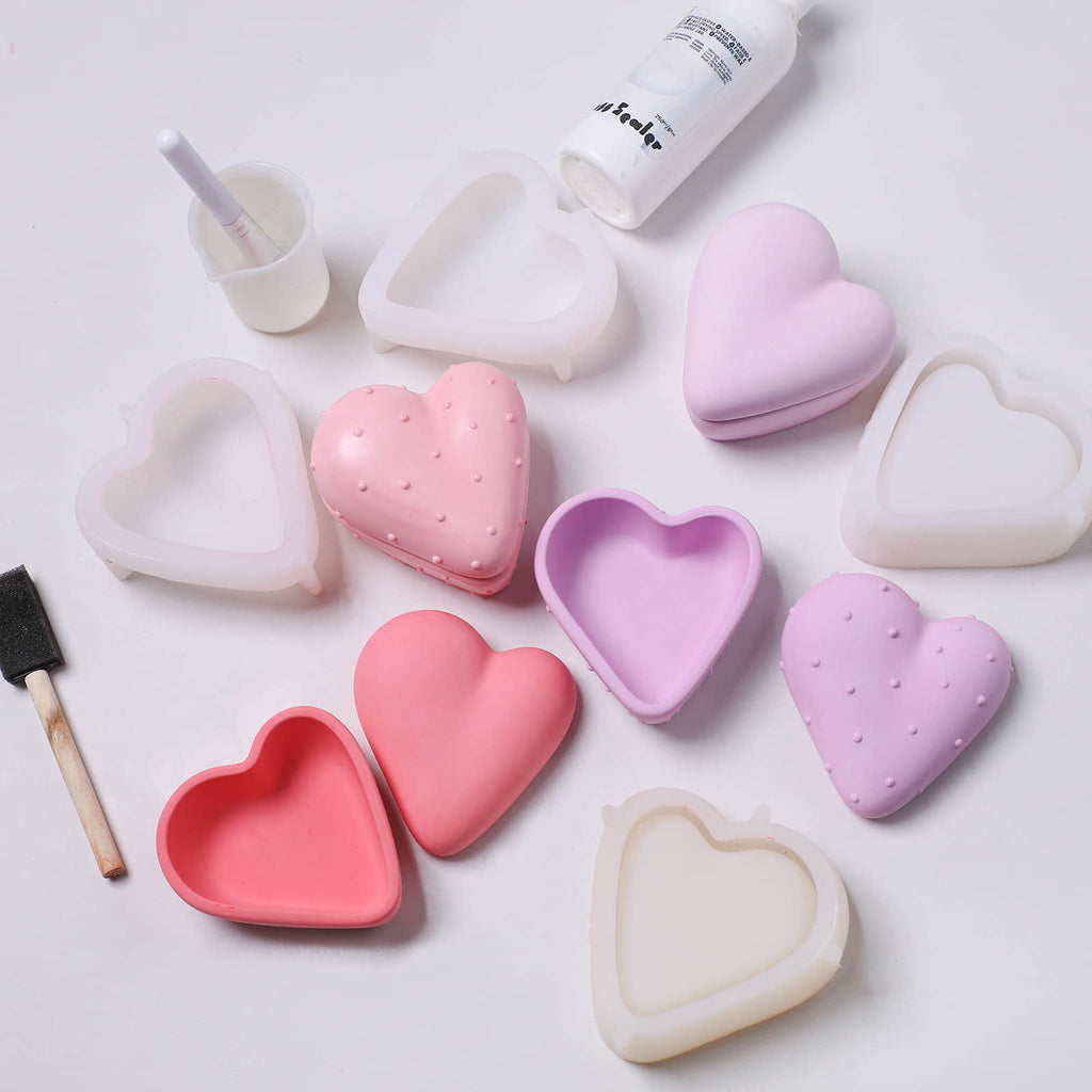 Pastel heart-shaped candle jar silicone molds and finished jars (smooth and dotted) with brush and sealer—DIY casting set on workspace - Boowan Nicole