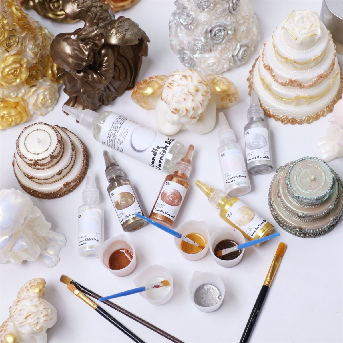 Candle gloss sealer bottles, mica paint cups and brushes arranged with gold and pearl tiered cake and angel candles on a white tabletop - Boowan Nicole