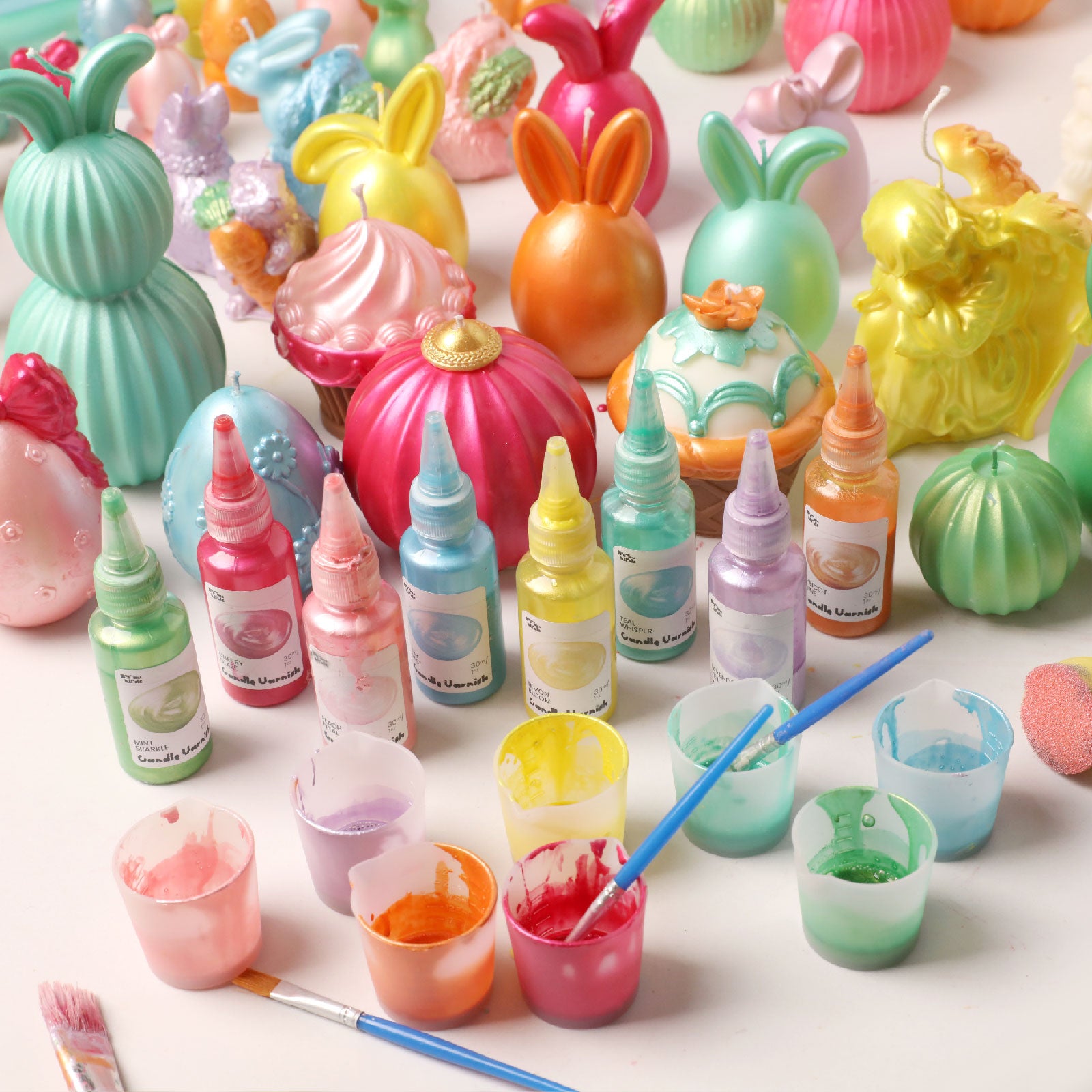 Colored candle varnishes and finished Easter-themed crafts arranged on a white table -Boowan Nicole