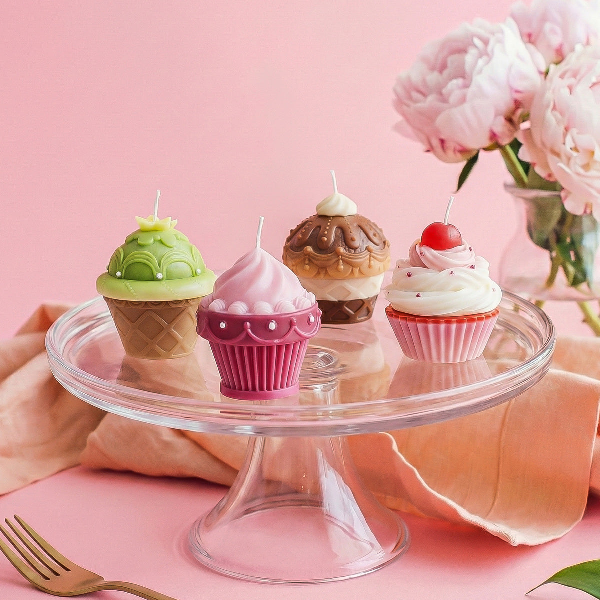 Four decorative dessert candles displayed on a glass cake stand with pink peonies against a pastel pink backdrop - Boowan Nicole