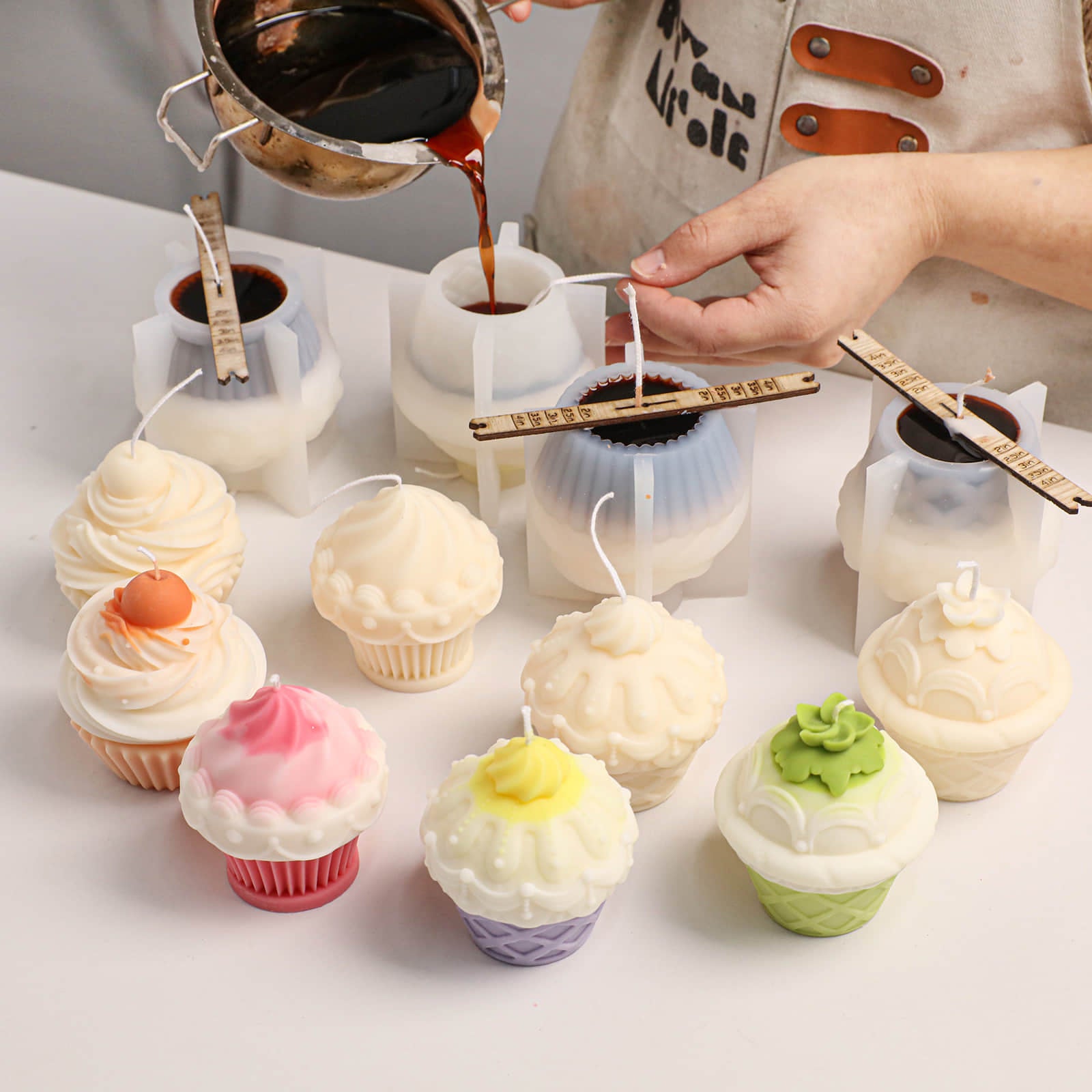 Artisan pouring melted wax into silicone molds, with finished white and pink cupcake candles on the workbench - Boowan Nicole
