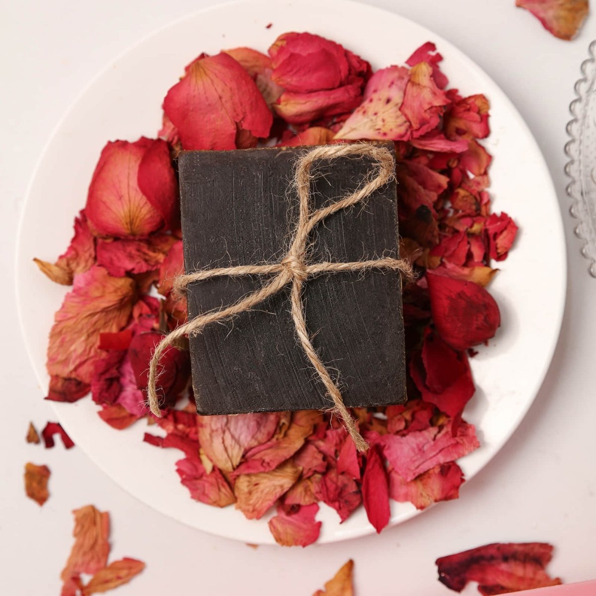 A dark square Damask Rose Essential Oil Soap Bar tied with rustic twine, elegantly presented on a bed of dried red rose petals - Boowan Nicole