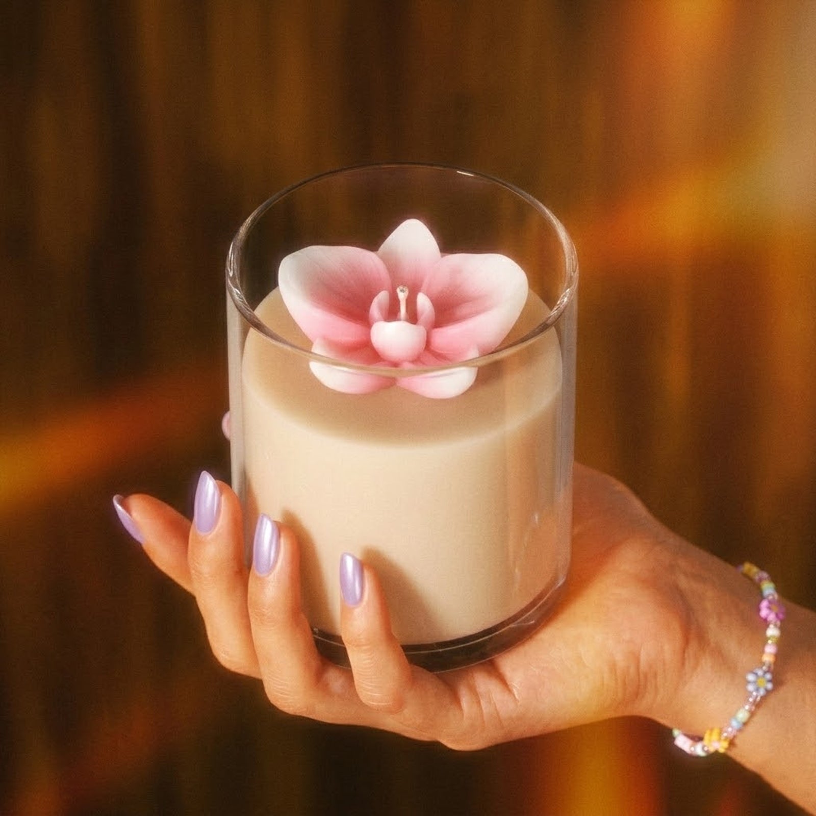 A hand holding a glass candle topped with a lifelike pink orchid wax topper, highlighting the mold's precise floral design. -Boowan Nicole