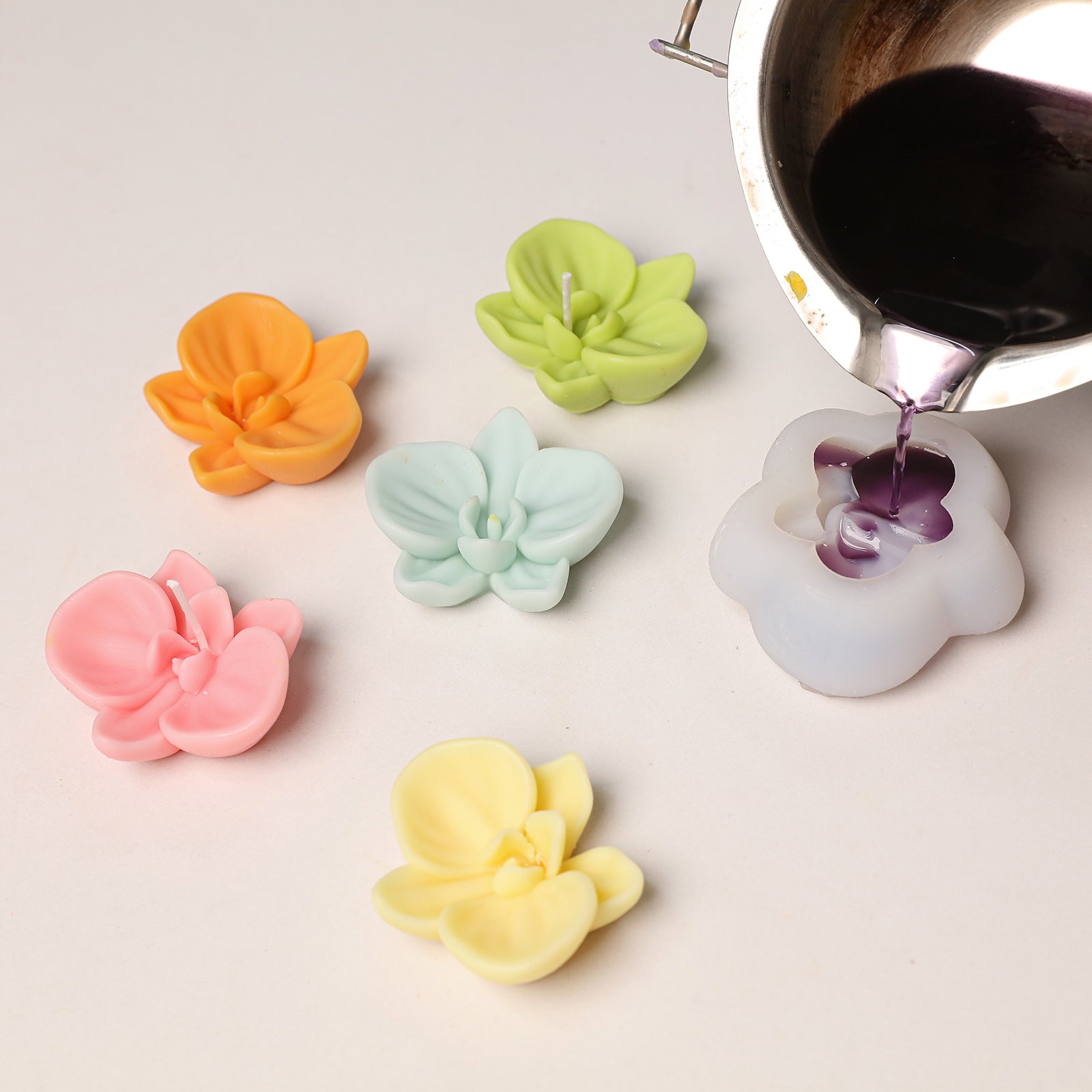 Colorful 3D orchid wax toppers in pink, orange, green, mint, and yellow, alongside a silicone mold being poured with wax. -Boowan Nicole