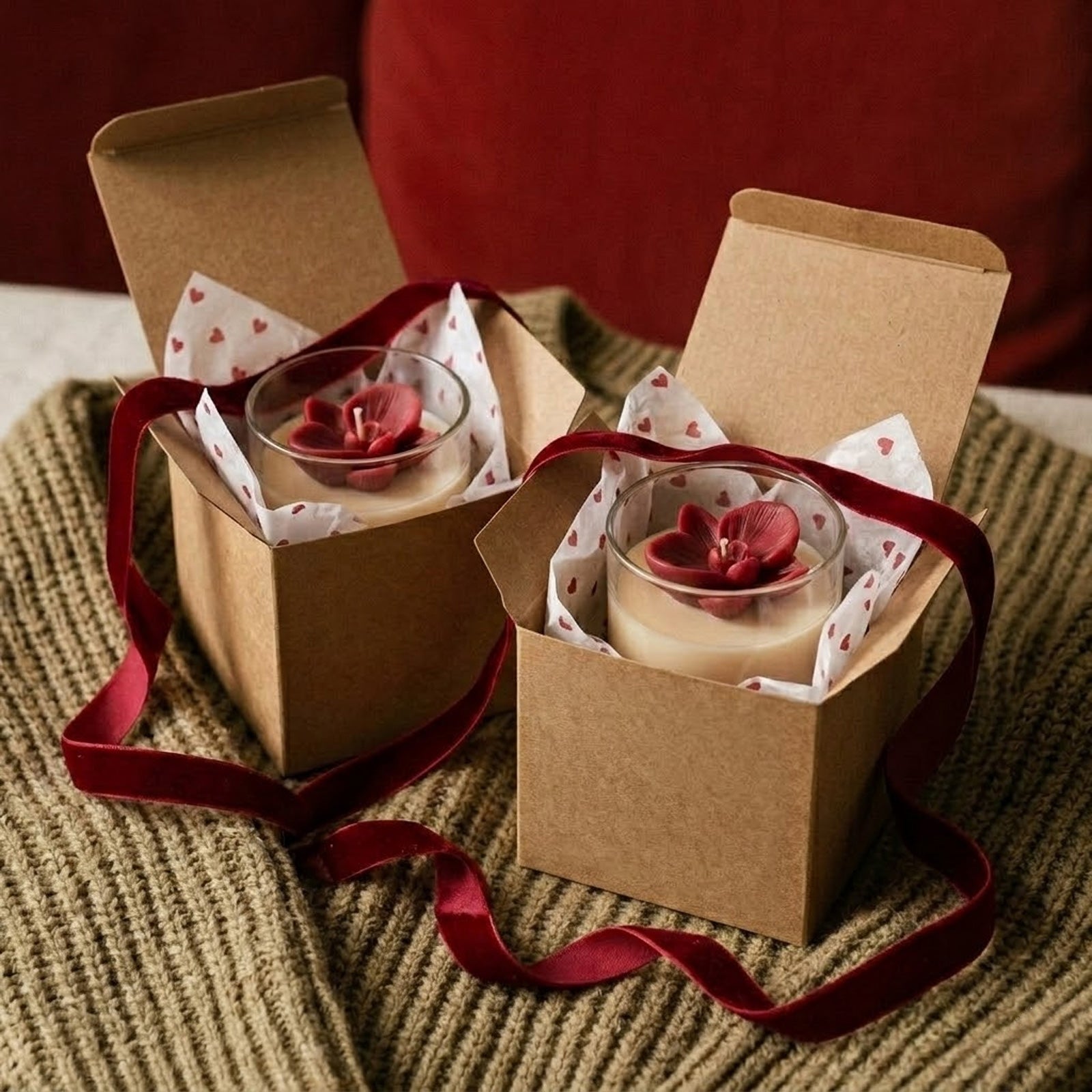 Two gift-boxed candles with red orchid toppers, wrapped in red velvet ribbon and heart-patterned tissue paper, ideal for gifting. -Boowan Nicole