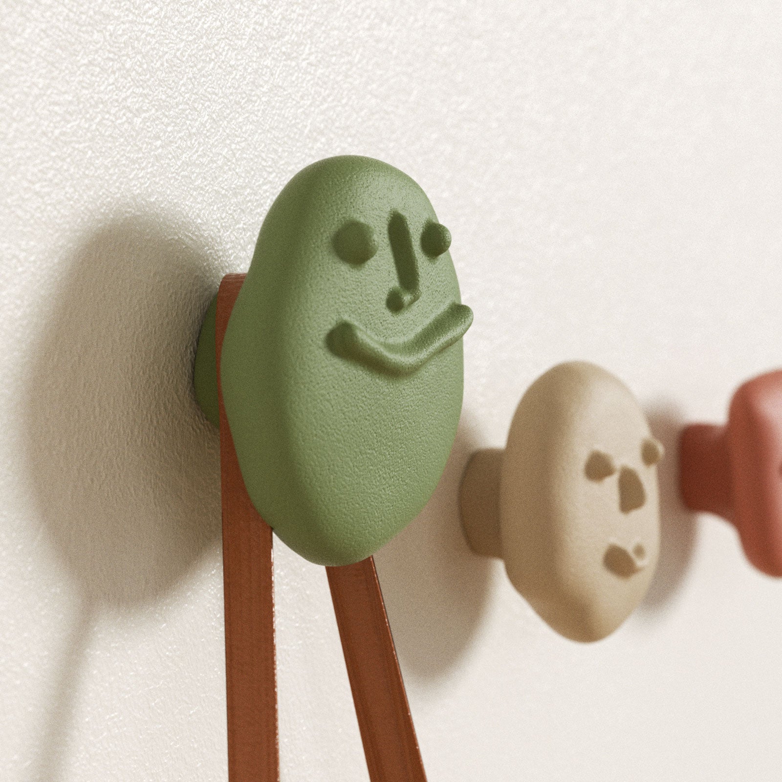 Green smiling pebble wall hook supporting bag straps, with beige and terracotta hooks in the background - Boowan Nicole