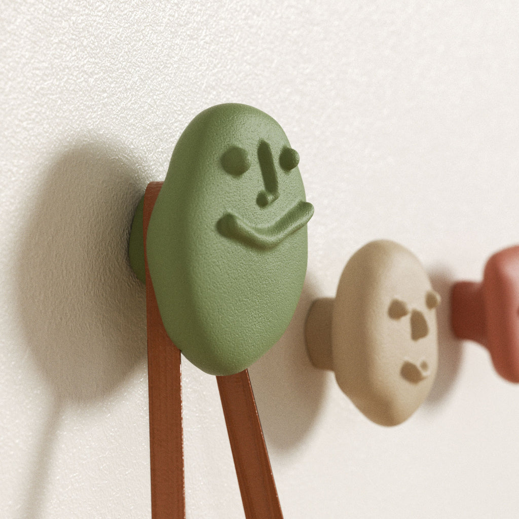 Green smiling pebble wall hook supporting bag straps, with beige and terracotta hooks in the background - Boowan Nicole