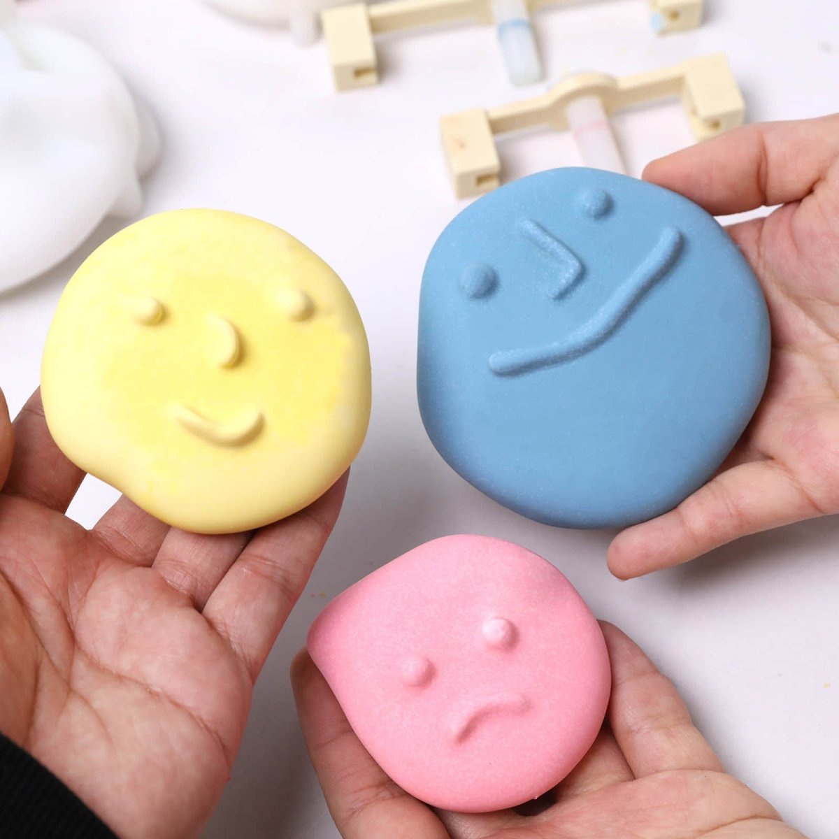 Hands holding three pastel pebble-face wall-hook casts—large blue smiling, medium yellow calm - Boowan Nicole