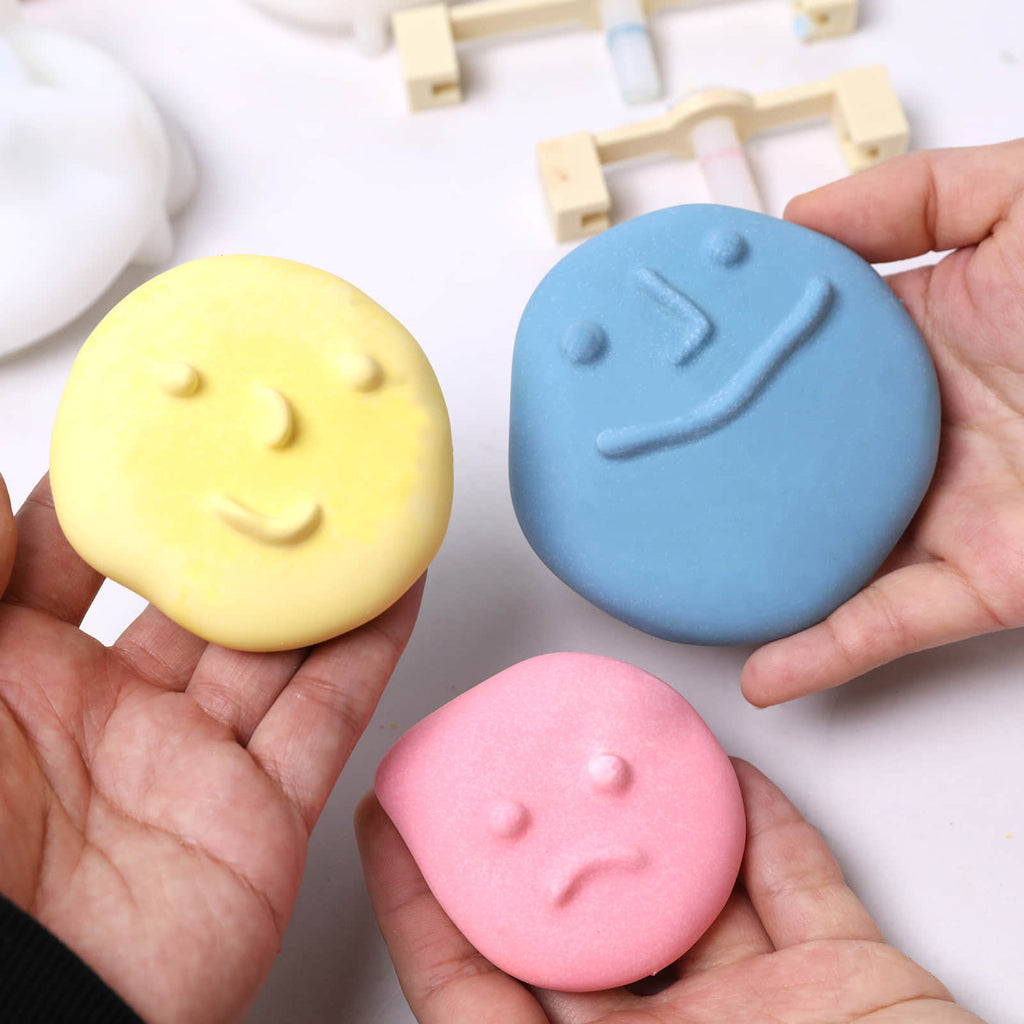 Hands holding three pastel pebble-face wall-hook casts—large blue smiling, medium yellow calm - Boowan Nicole